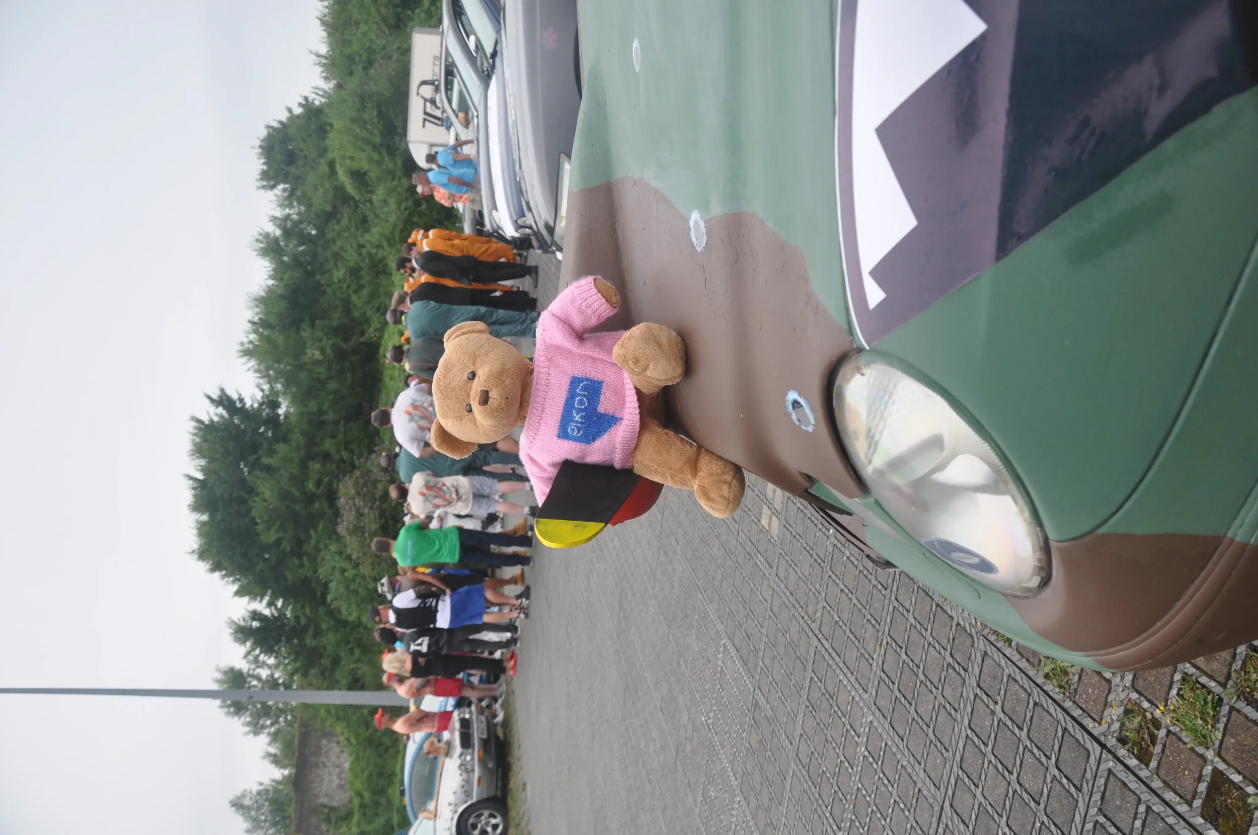 A stuffed teddy bear wearing a pink sweater with the word 'OK' on it, sitting on the hood of a green car. In the background, a group of people are in line at a car show or gathering, surrounded by trees.