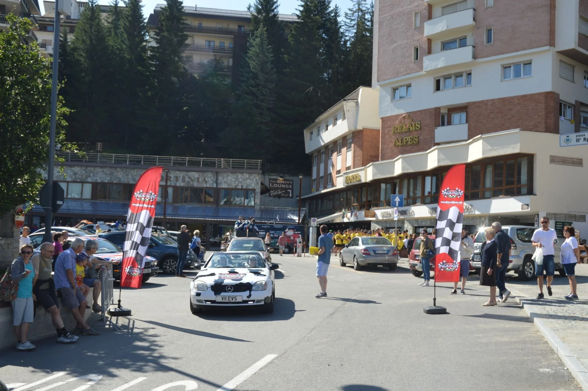 People gathering in a parking lot during a car event, with a white convertible car in front, flags on either side, and buildings with signs in the background.