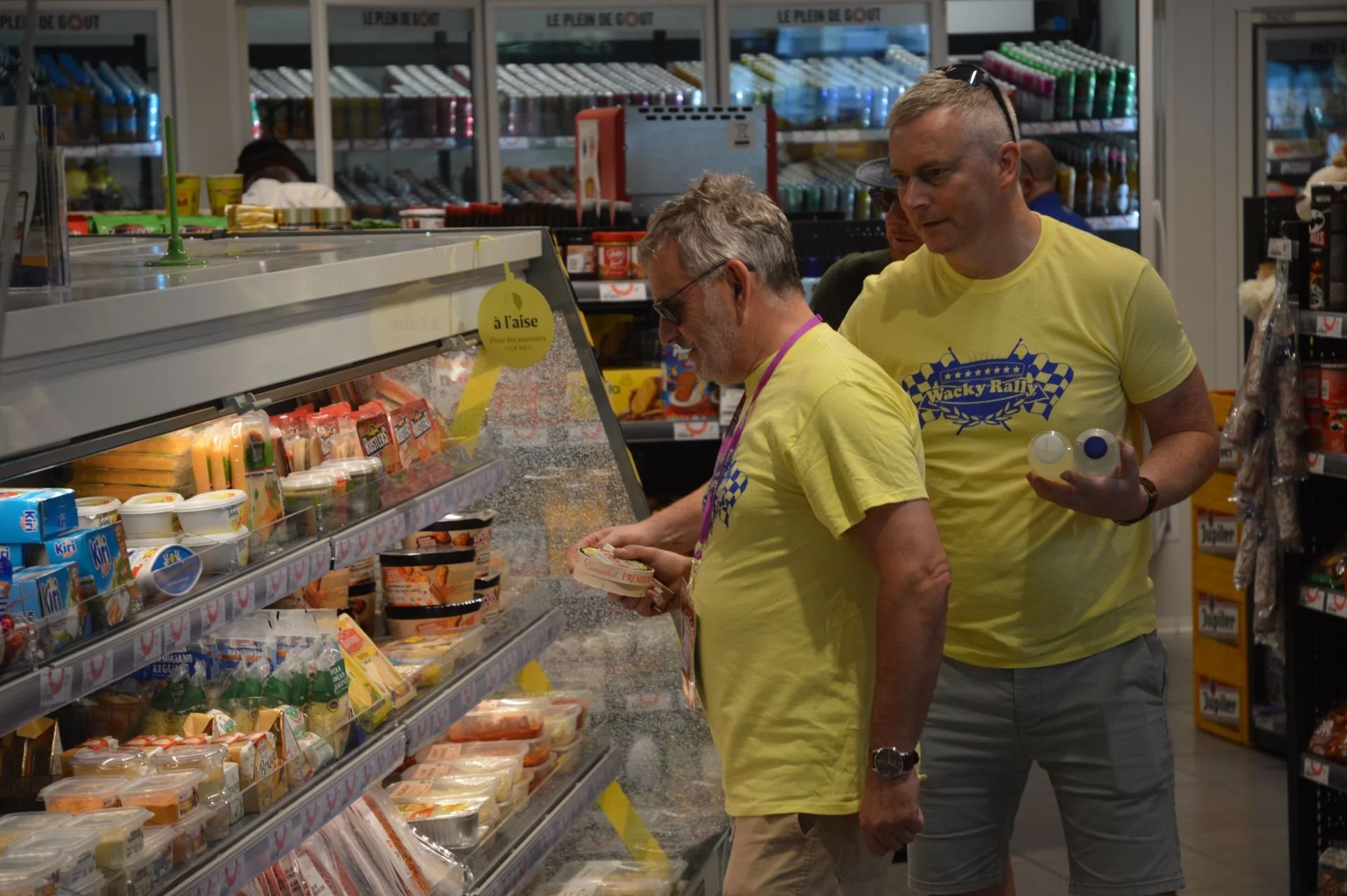 Two men in yellow t-shirts shopping for cheese in a grocery store. One is holding a pack of cheese, the other is holding bottles, and they are standing near refrigerated shelves with various dairy products.