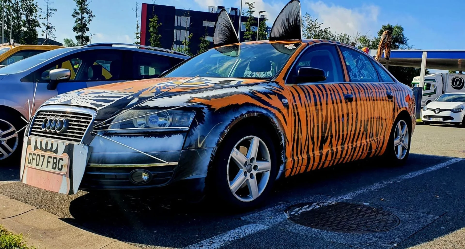 A car decorated with tiger stripes, featuring 3D tiger ears on the roof and a tiger face painted on the hood, parked in a parking lot.