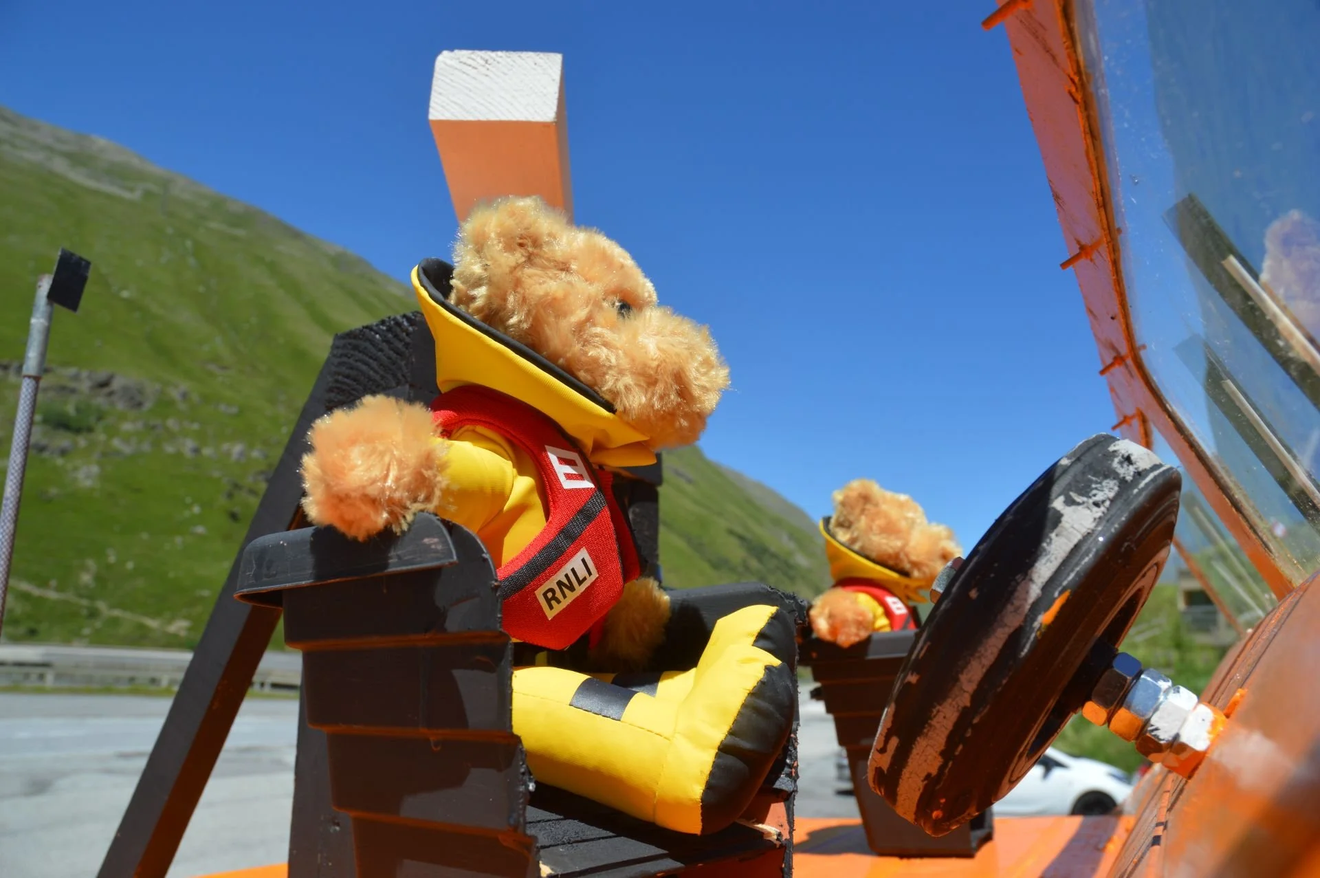 Two stuffed teddy bears dressed as rescue workers sitting in the cockpit of an orange rescue vehicle, with a mountain and blue sky in the background.