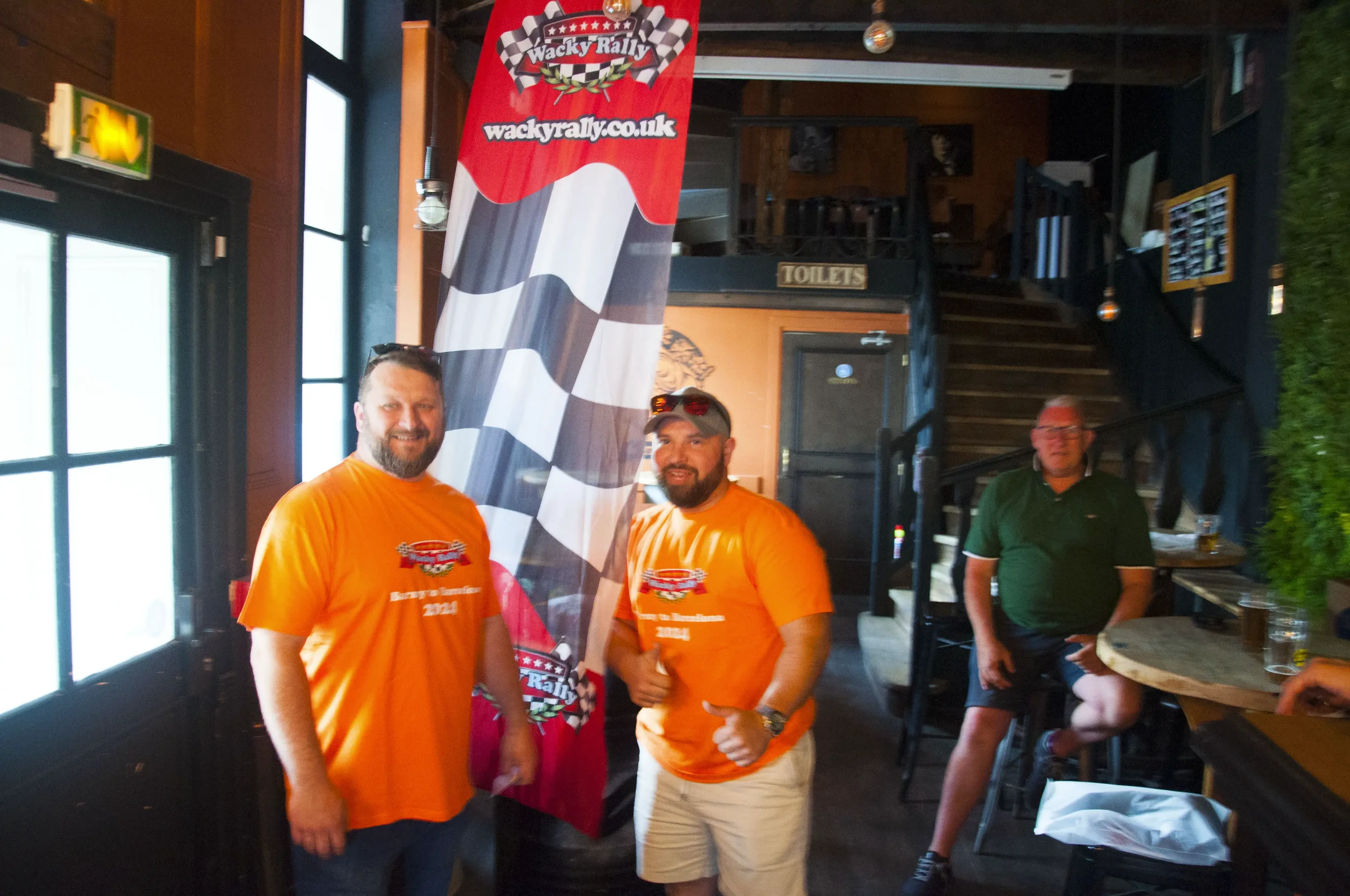 Three men inside a pub or restaurant, two wearing bright orange T-shirts with a rally logo, standing near a tall flag with a checkered pattern and rally branding. Another man in a green polo and shorts is sitting at a high table in the background nea
