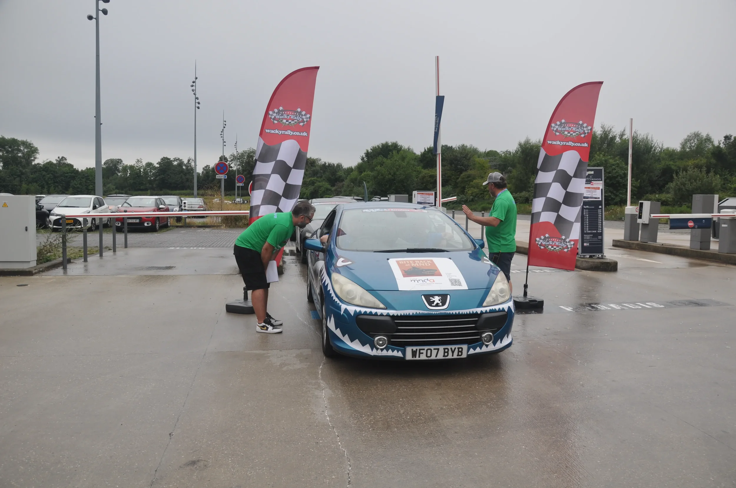 A blue Peugeot car with shark teeth design on the front is at a checkpoint with two men in green shirts talking to the driver. Red checkered flags with 'Wacky Rallies' logos are displayed.