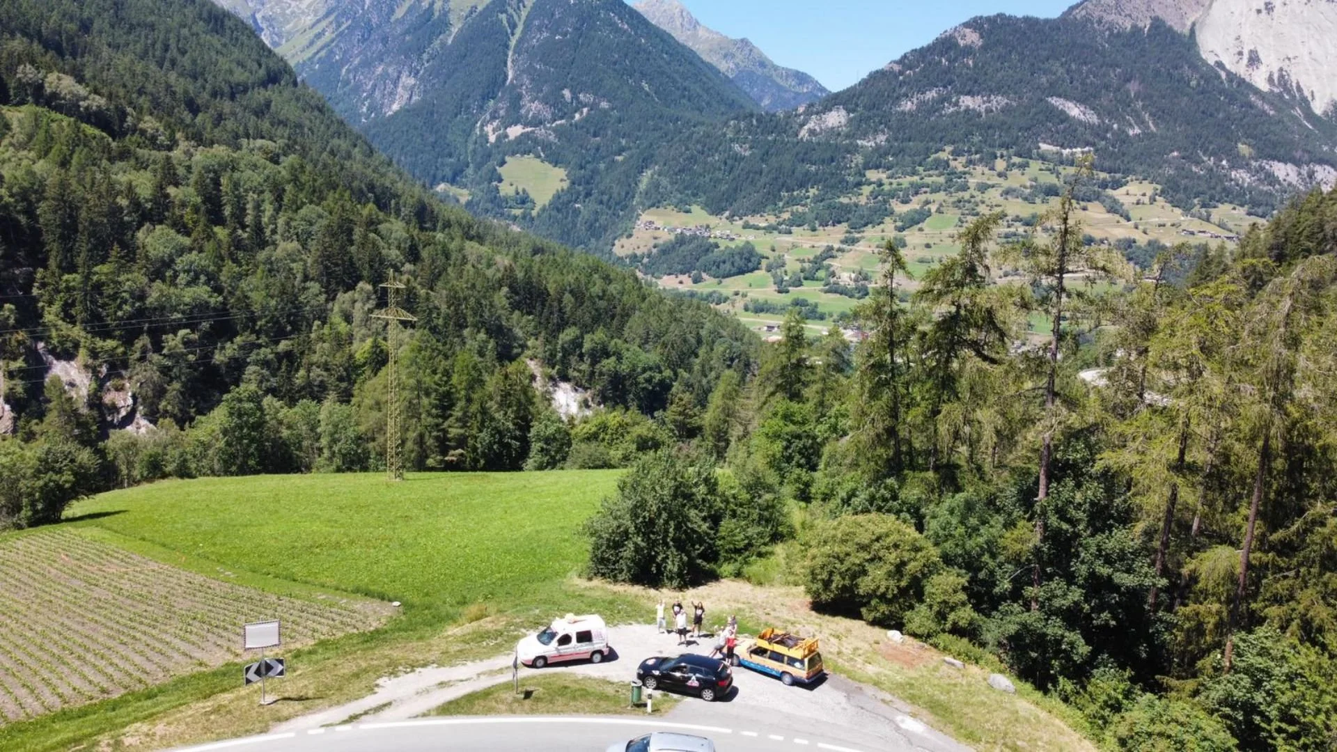 A scenic mountain landscape with lush green trees, grassy fields, and steep mountains in the background. At the bottom, there is a small parking area with a few cars and a group of people standing near a yellow vehicle.