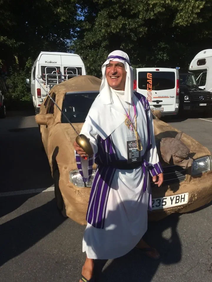 A man dressed in Middle Eastern attire standing in front of a car that looks like it is made of paper or cardboard, with other vehicles in the background.