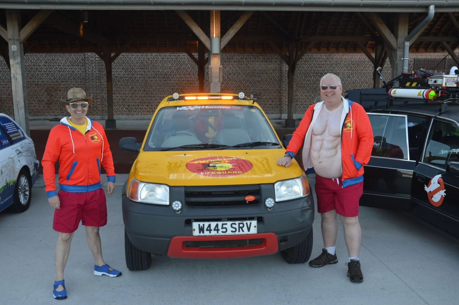 Two men in red jackets and shorts pose with a yellow lifeguard vehicle featuring a logo and license plate, under a covered area near other themed cars.