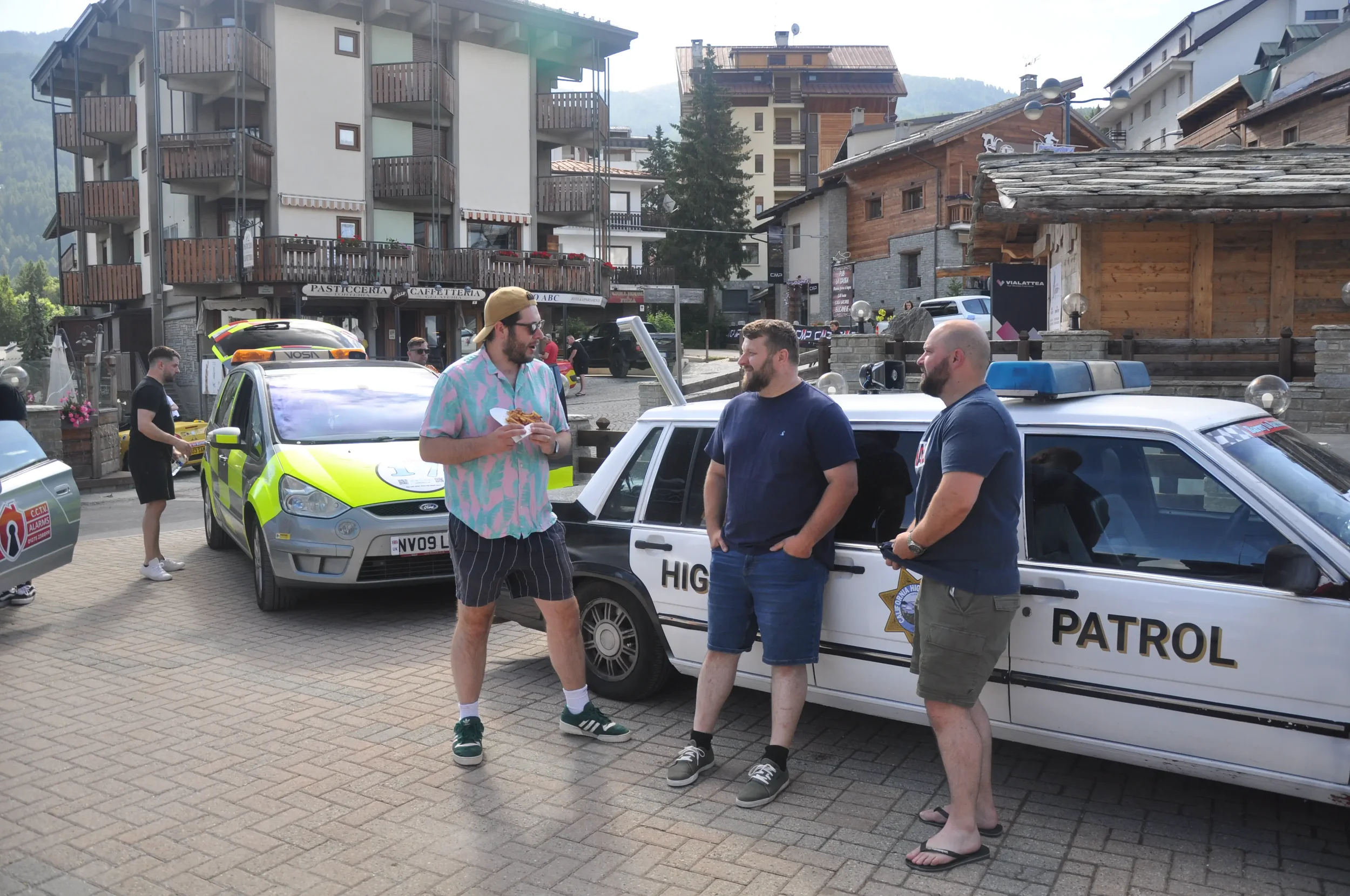 Three men standing and chatting near a police car, with another police car behind them. One man is eating food. The background features buildings, a city square, and a mountain.