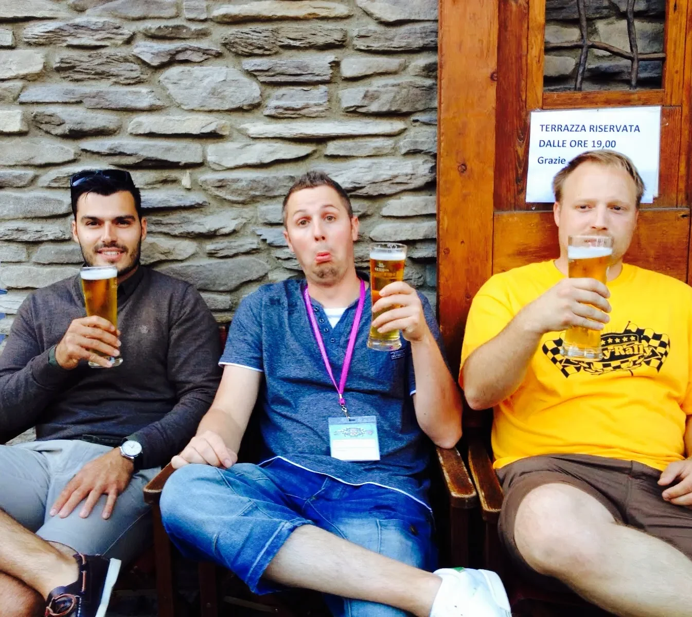 Three men sitting together holding glasses of beer, sitting on a bench against a stone wall with a wooden panel and a sign in Italian behind them.