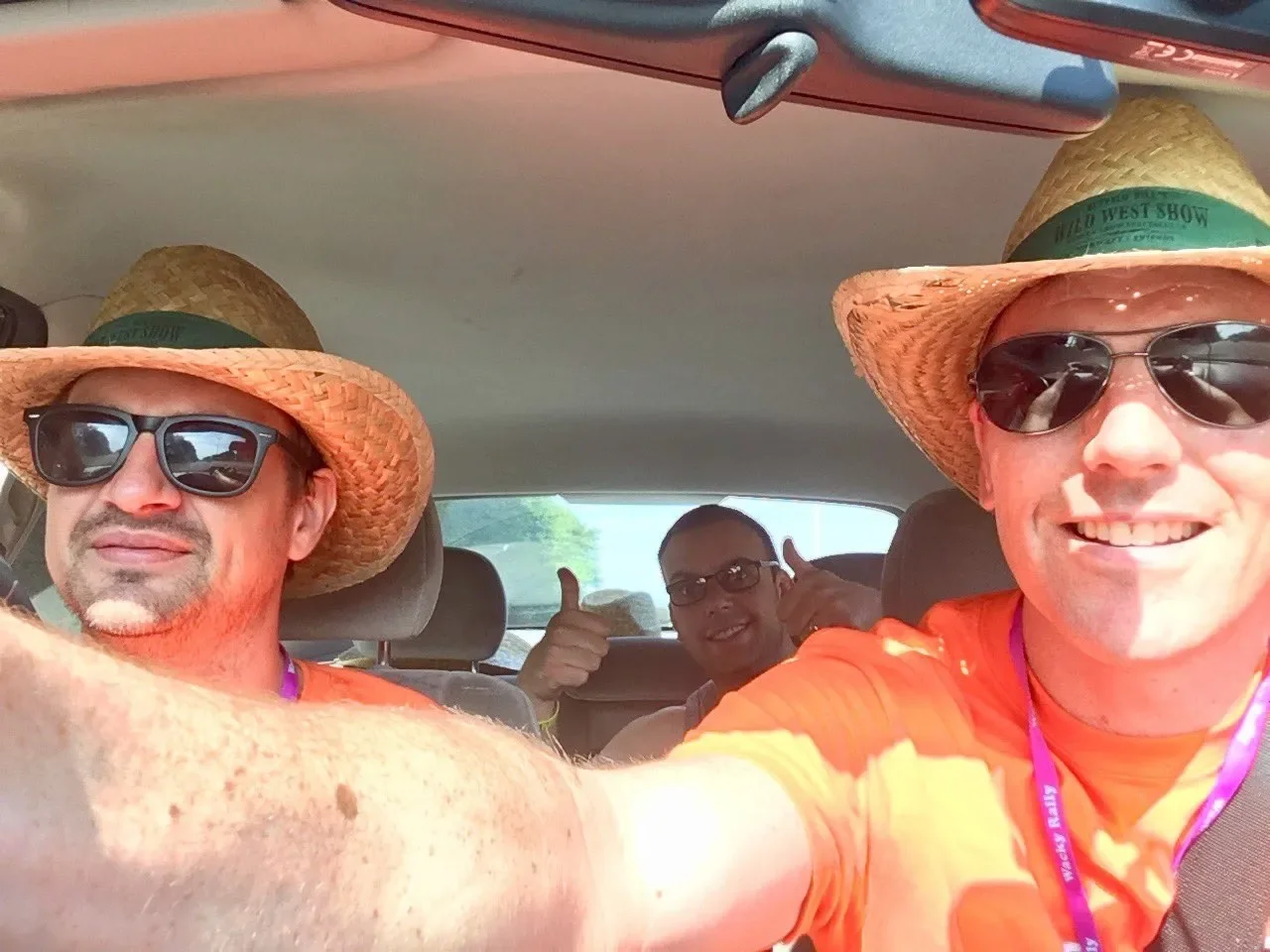 Four men inside a car. They are wearing straw hats and sunglasses. The man in front on the right is taking a selfie, smiling. The man on the left is also smiling, and the two men in the back are giving thumbs-up signs, smiling. The men are dressed in