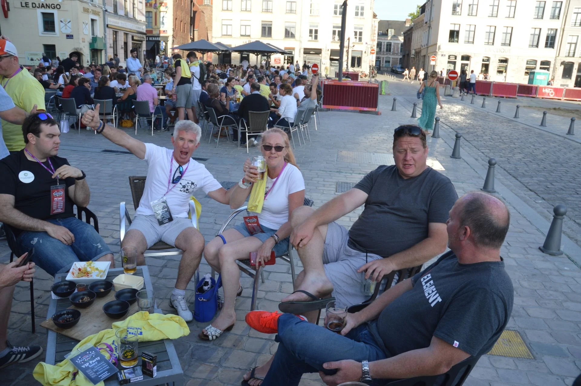 Group of five friends sitting outdoors at a social gathering in a city square, with some holding drinks and engaging in conversation, others eating food, and a large crowd in the background at outdoor tables.