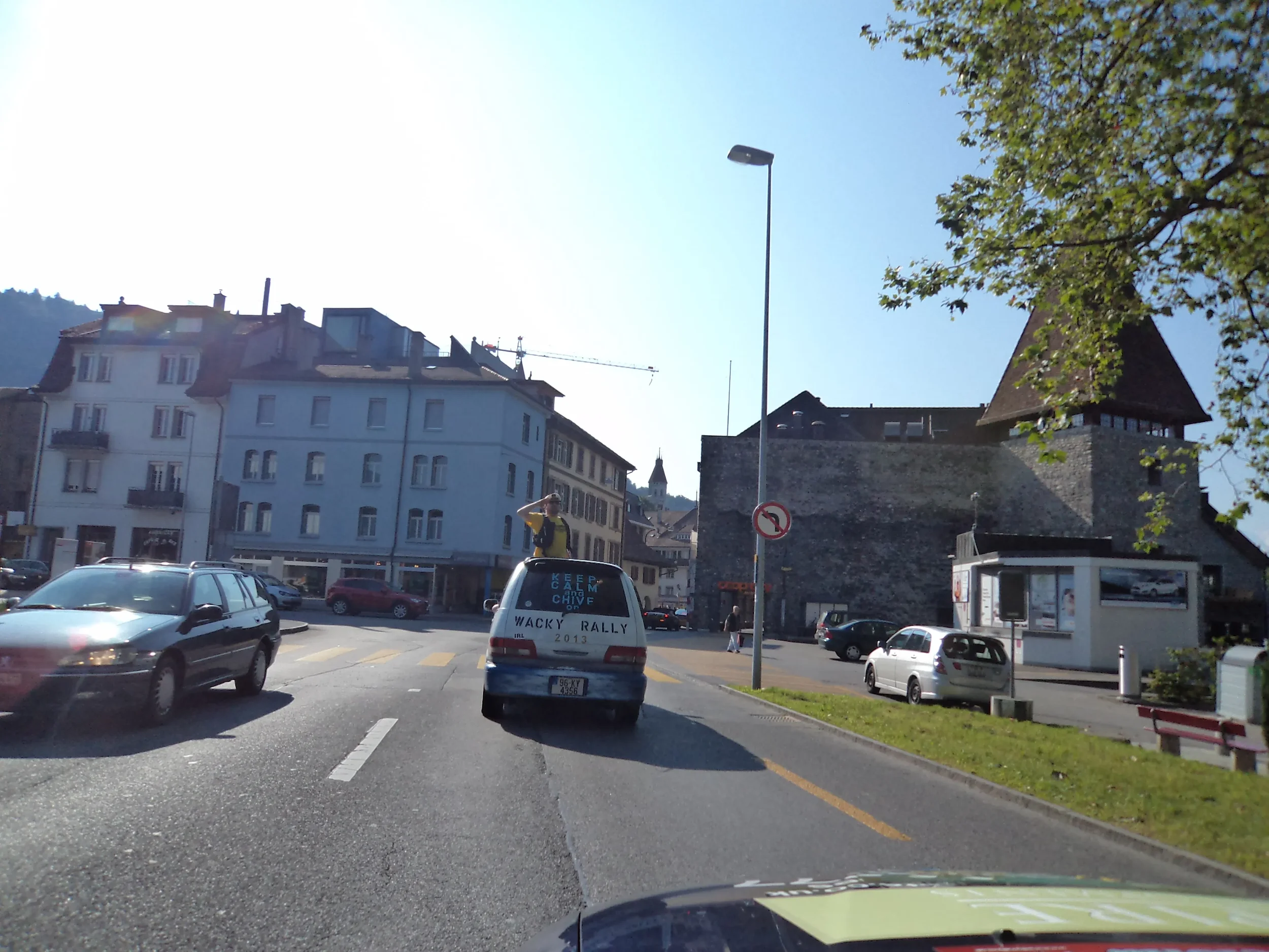 Street view with cars, a person in a yellow jacket standing in the back of a vehicle, historic stone building with a tower, and modern buildings in a European town.
