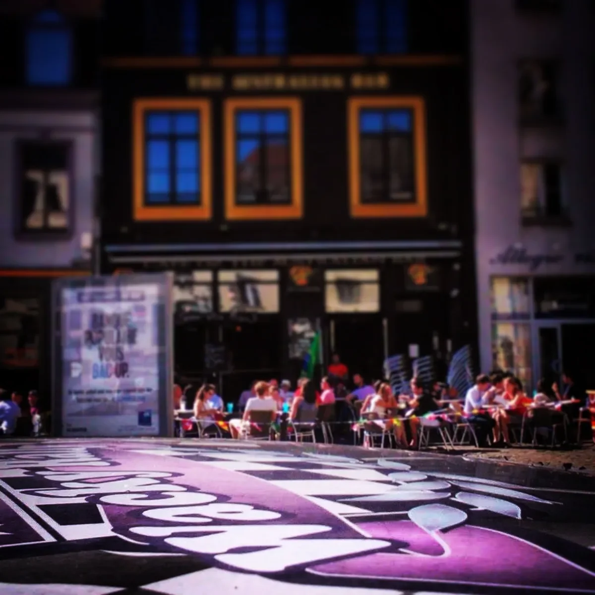 People dining at outdoor tables in front of a black building with illuminated windows, with a large painted sign on the street in the foreground.