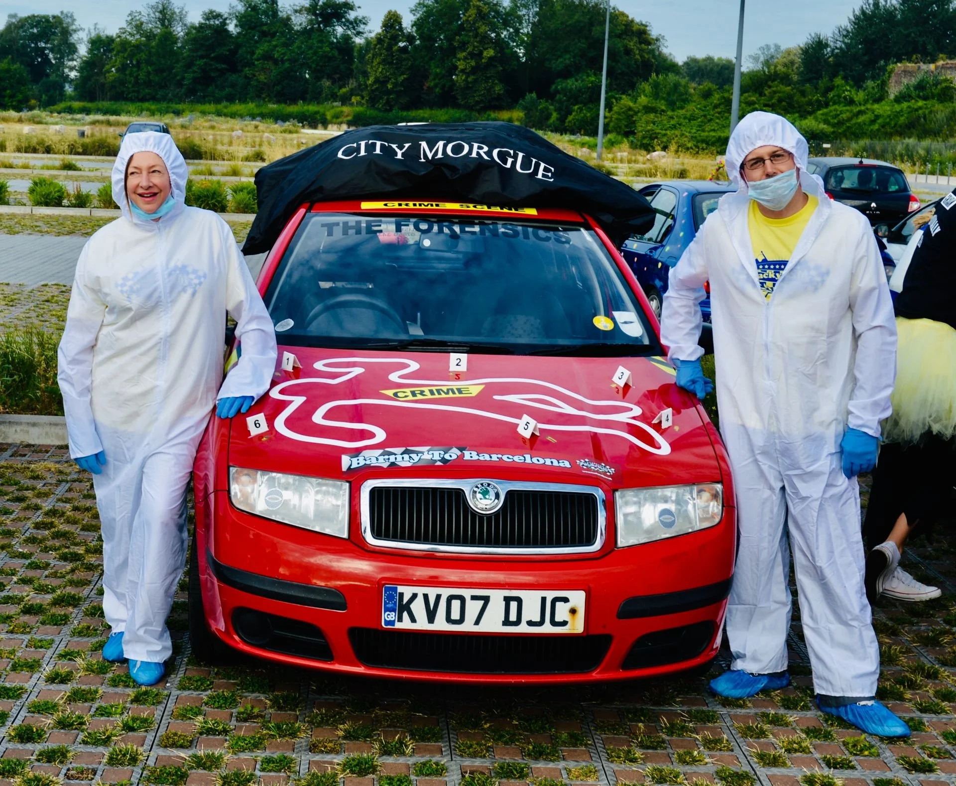 Two people dressed as forensic investigators in white coveralls, blue gloves, and shoe covers, standing next to a red car with crime scene markings and labels, in an outdoor parking area.