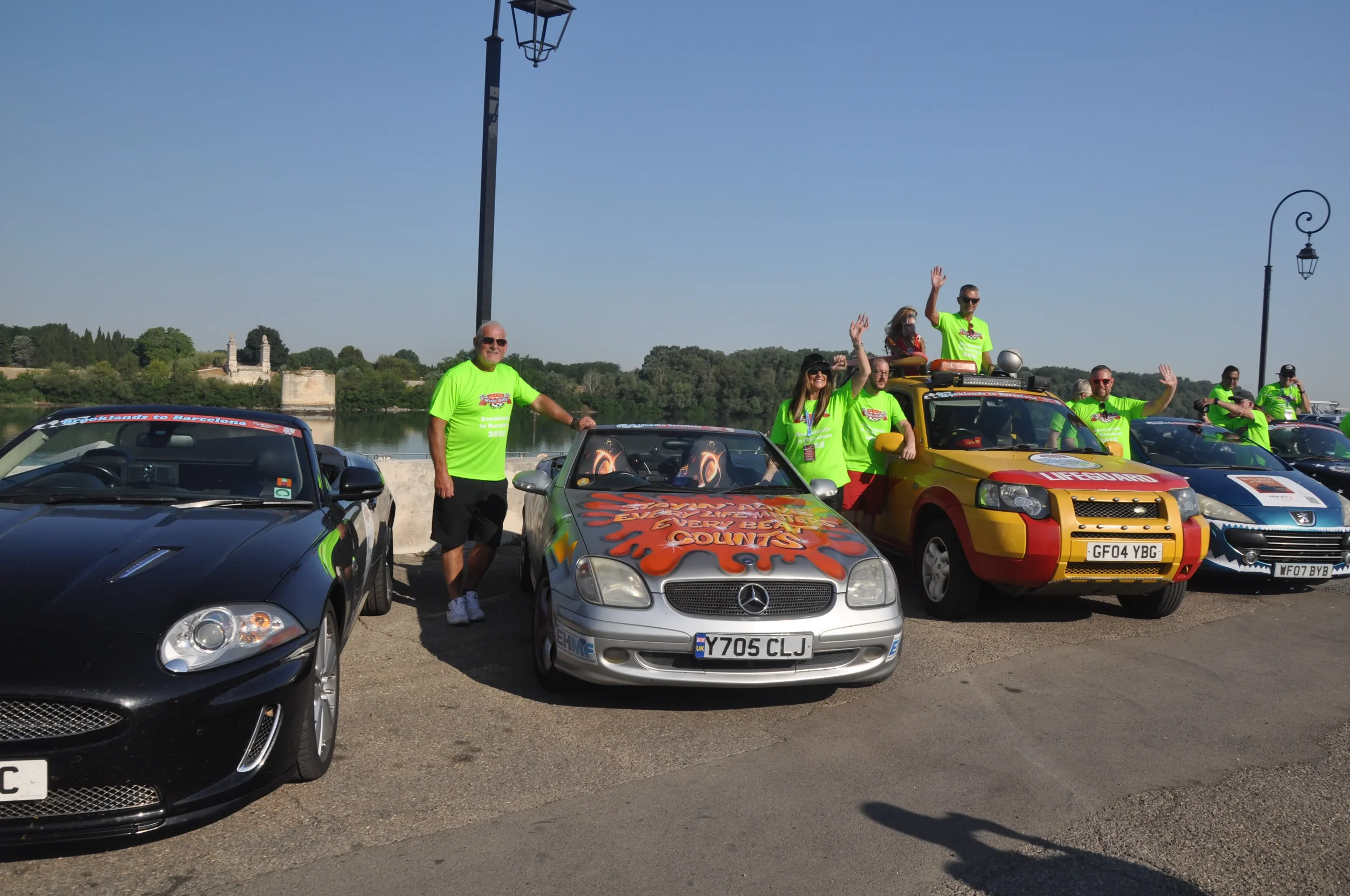 Group of people in bright green shirts standing next to decorated cars, waving and smiling during a sunny outdoor event by a lake.