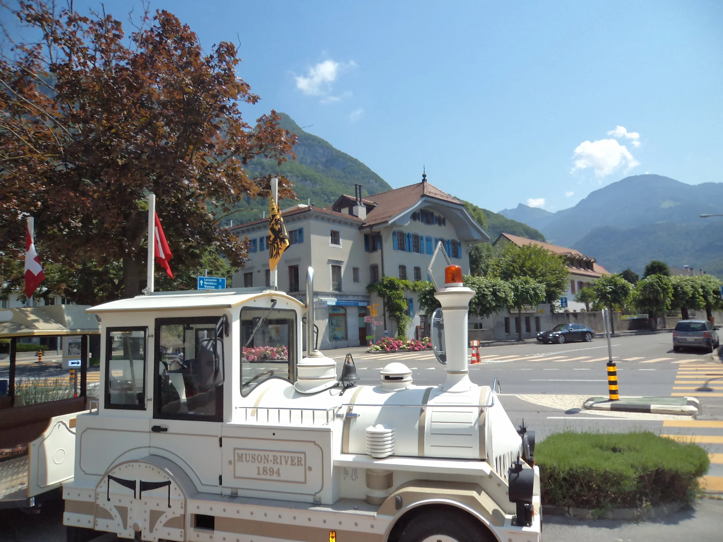 A miniature train with the words "Muson River 1894" parked on a street in a scenic mountain town with buildings, trees, and mountains in the background.