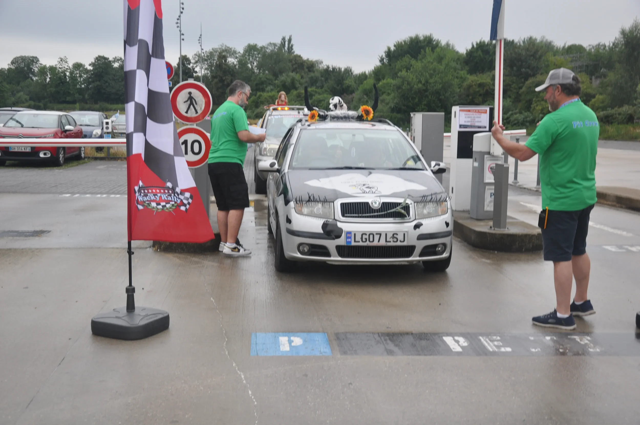 A gathering at a parking lot checkpoint with two men in green shirts and a car decorated with a dog and sunflowers on the roof. One man is standing at a barrier gate, the other by the car, which has a license plate ending in L3J. A checkered flag ban