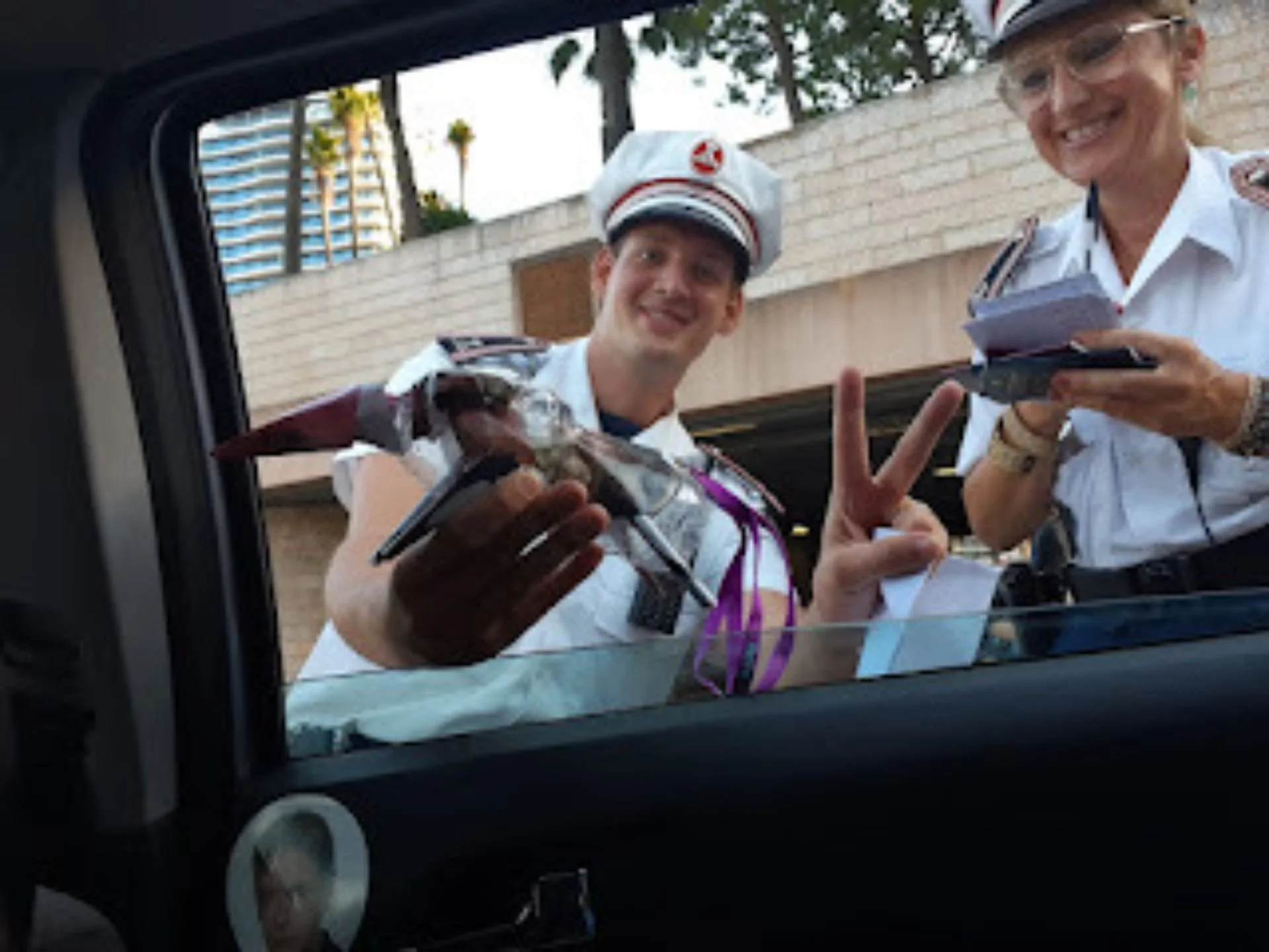 Two people dressed as police officers standing outside, smiling, with one making a peace sign. Their uniform hats feature a badge, and they are holding clipboards and a model police car. The background includes a brick wall, some trees, and a tall bu