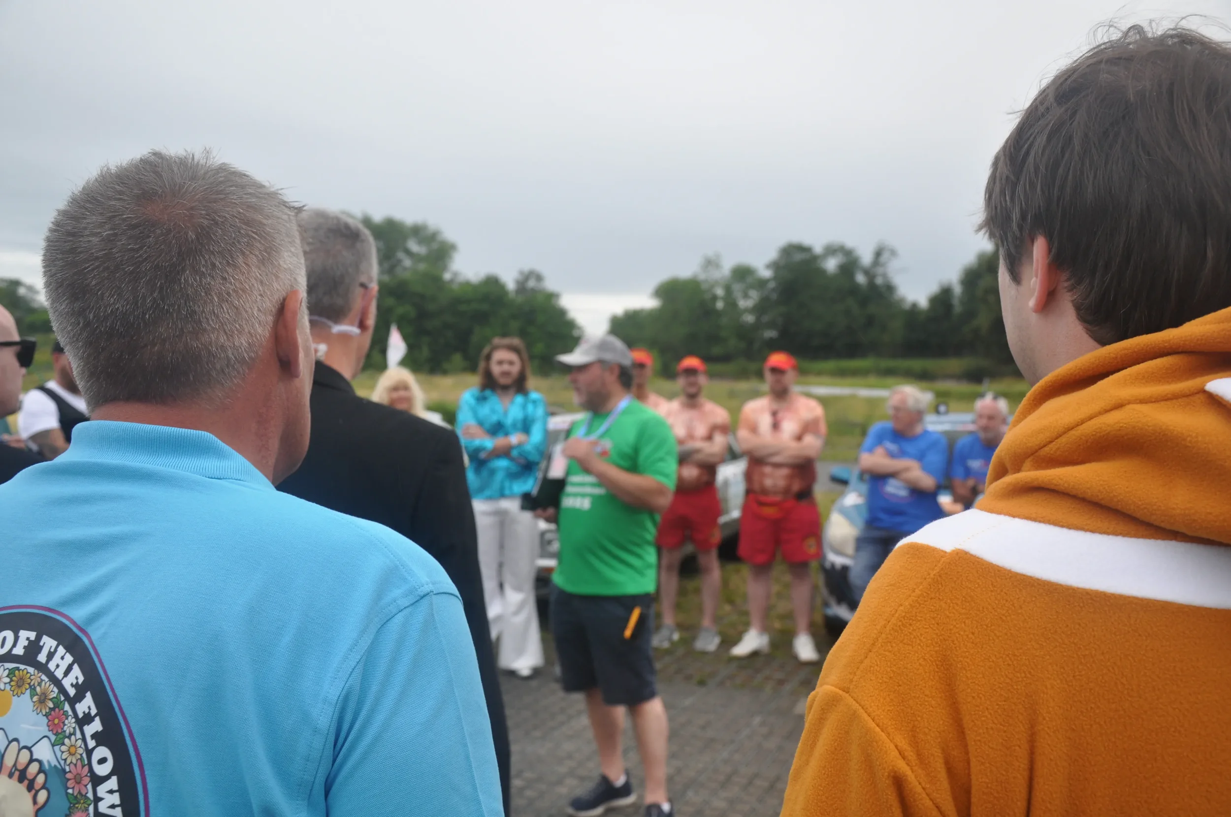 Group of people outdoors, some in casual clothing and some in uniforms, gathered around a man speaking, with a background of trees and overcast sky.