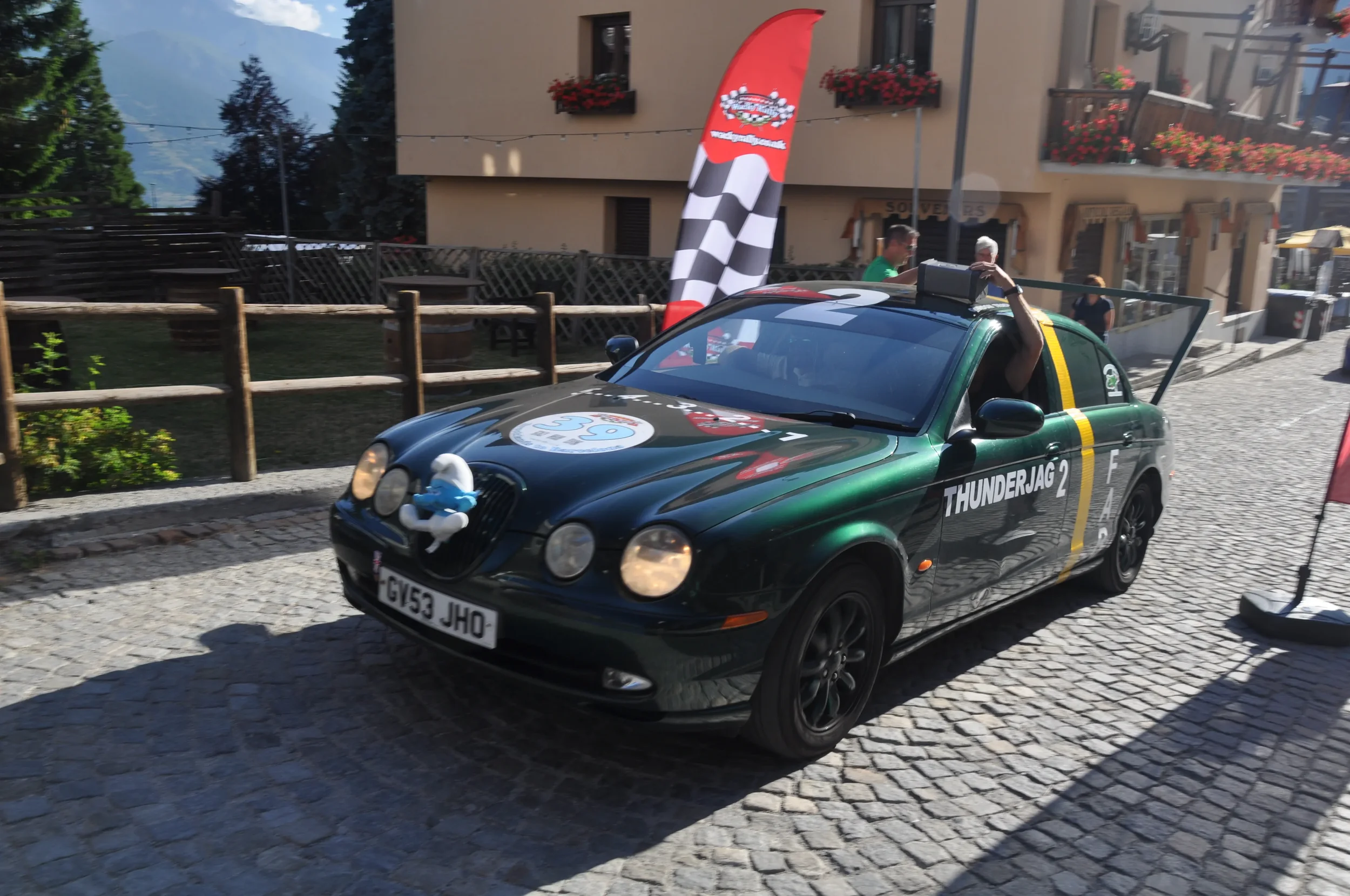 A black convertible car with racing decals and the label 'THUNDERJAG 2' parked on a cobblestone street. The car has a large rear spoiler, racing stripes, and a plush snowman figure attached to the front grille. A person is waving from the passenger s