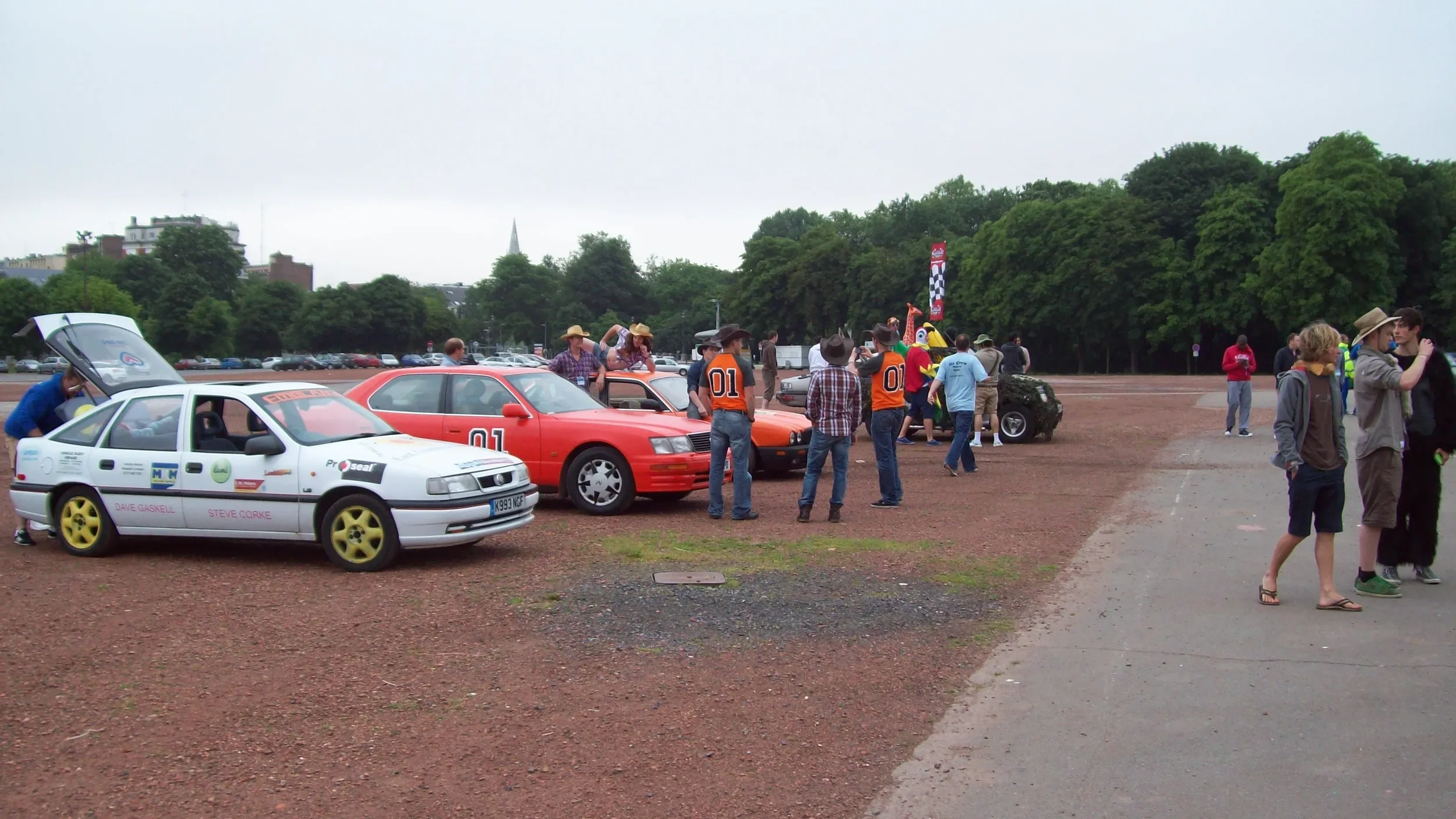 Group of people at a car show with vintage cars, some people in cowboy hats, and a person in a colorful mascot costume, trees in the background, on an overcast day.