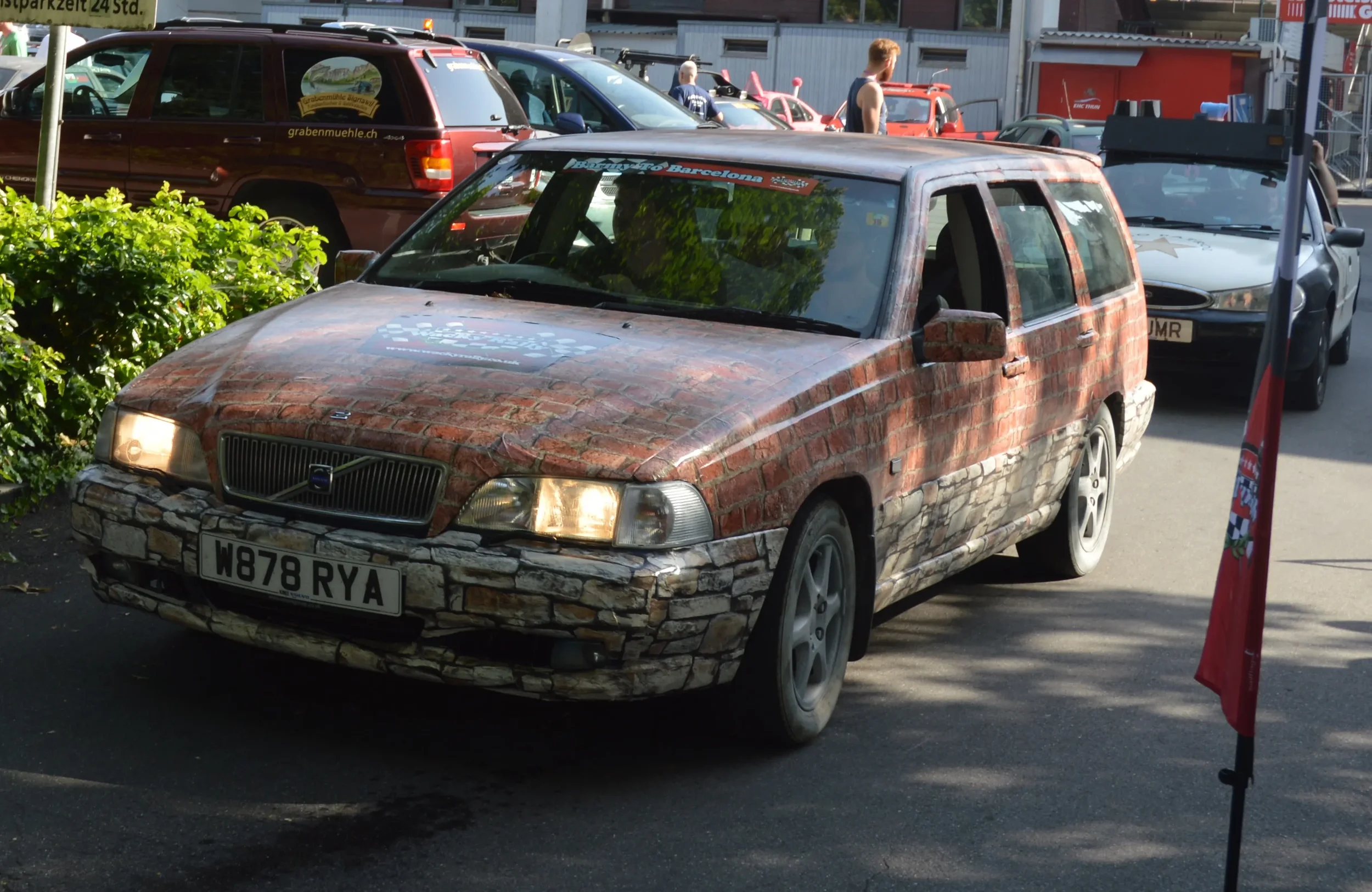 A car with a brick and stone wrap design, parked outdoors in a parking lot, with other vehicles and people in the background.