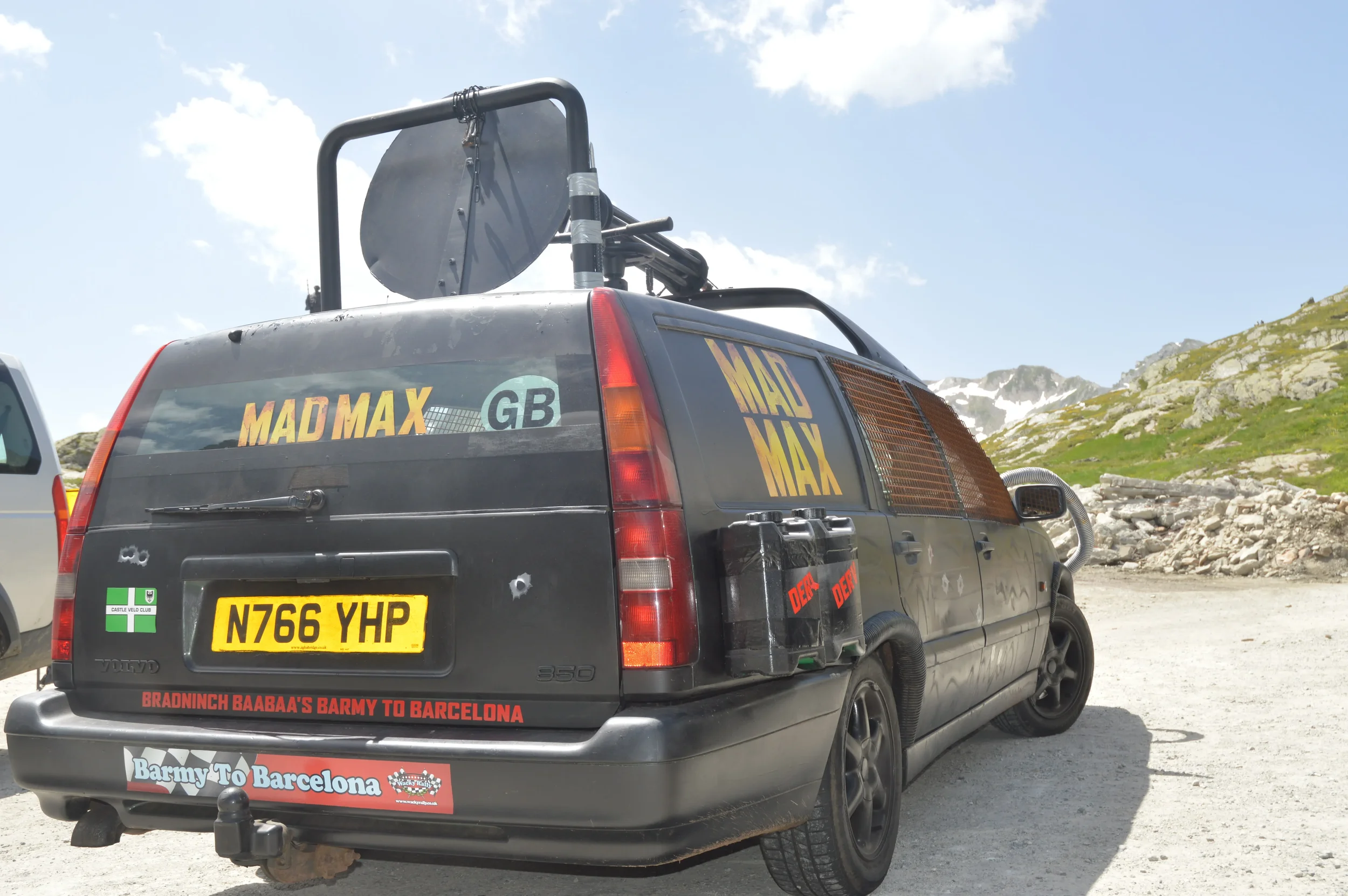 A black car with multiple stickers, including 'MAD MAX,' a license plate 'N766 YHP,' and 'Barmy To Barcelona,' is parked outdoors on a gravel area with mountains and green landscape in the background.