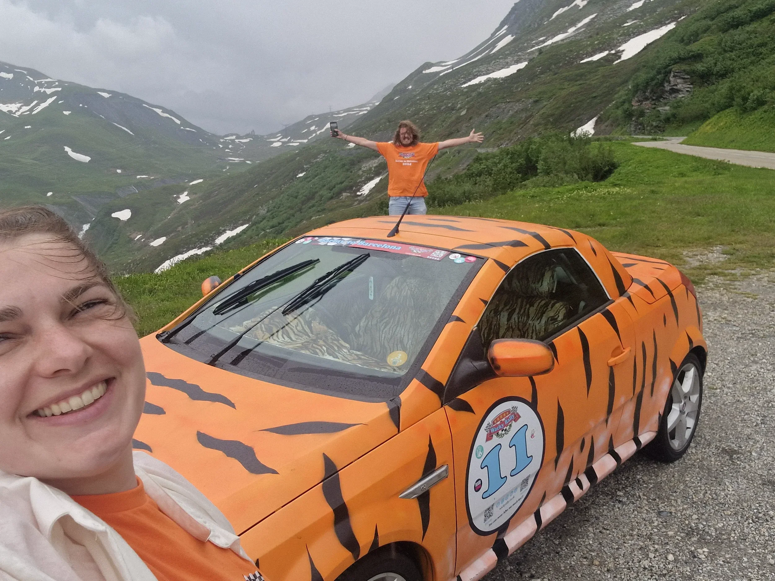 Two women taking a selfie in front of a tiger-striped orange car with a mountain landscape in the background. One woman is in the foreground smiling, and the other stands with arms outstretched on top of the car.