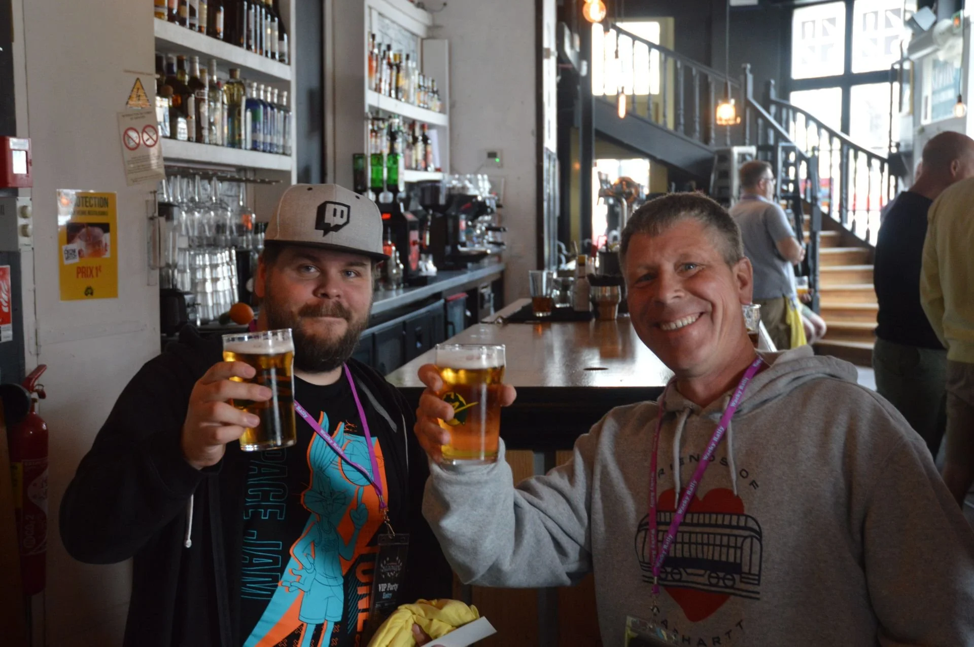 Two men smiling and raising glasses of beer inside a bar. The man on the left wears a gray cap and a black shirt with colorful graphics. The man on the right wears a gray hoodie and has a pink lanyard around his neck. The bar has bottles of alcohol a