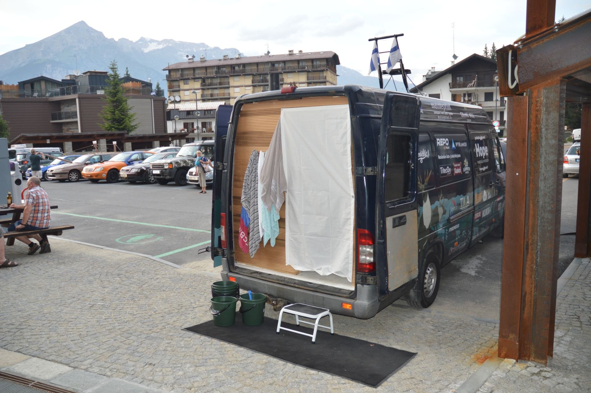 Mobile laundry or drying service van with hanging towels and clothes, buckets, and a step stool in front, parked outside with a mountain backdrop.