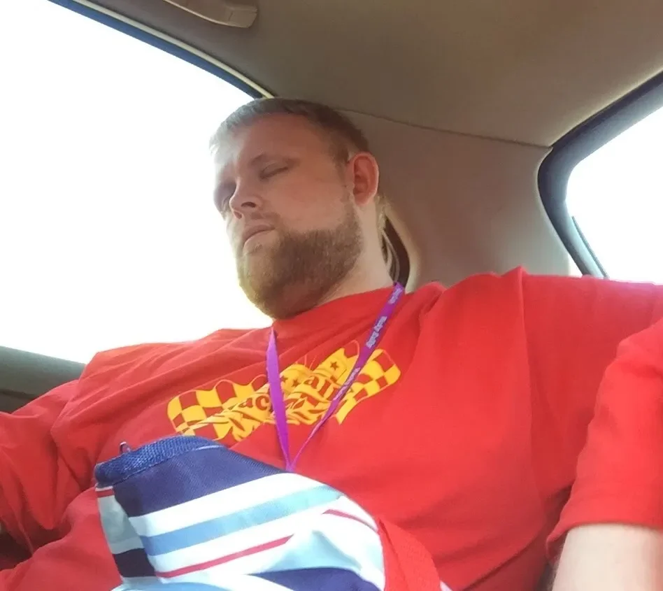 A man with a beard and long hair tied back, wearing a red shirt with a yellow and red design, sitting in the backseat of a vehicle with closed eyes, resting or sleeping.