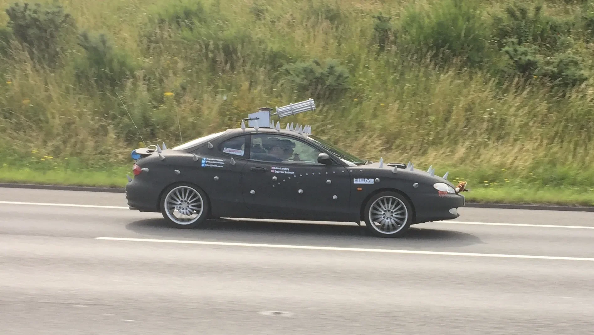 A black convertible car on a highway decorated with various spikes and props resembling a strange sci-fi or fantasy vehicle.