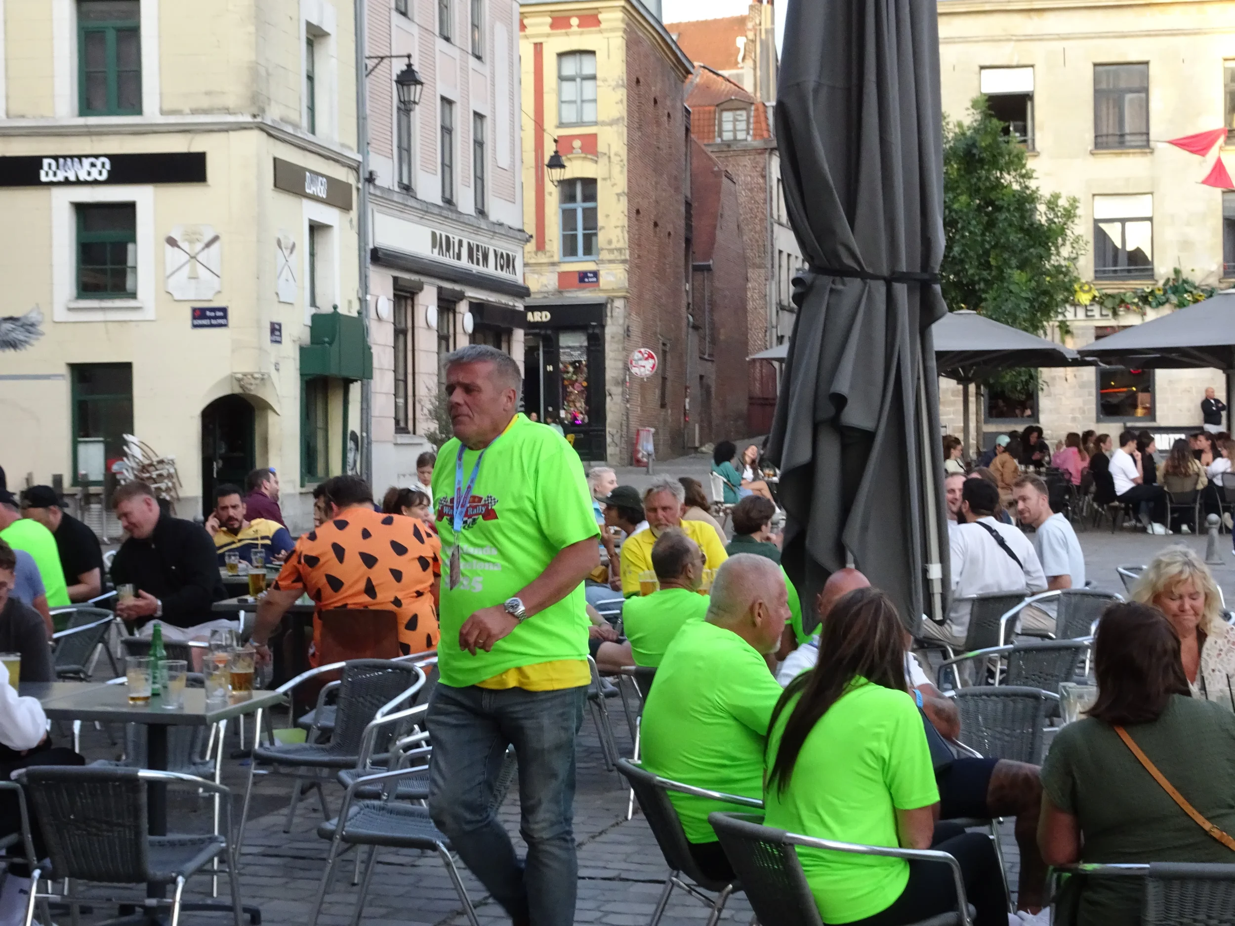 Crowd of people sitting at outdoor tables of a restaurant or cafe in a city square, with some wearing bright green shirts, and buildings with signs, including one that says 'PARIS NEW YORK', in the background.