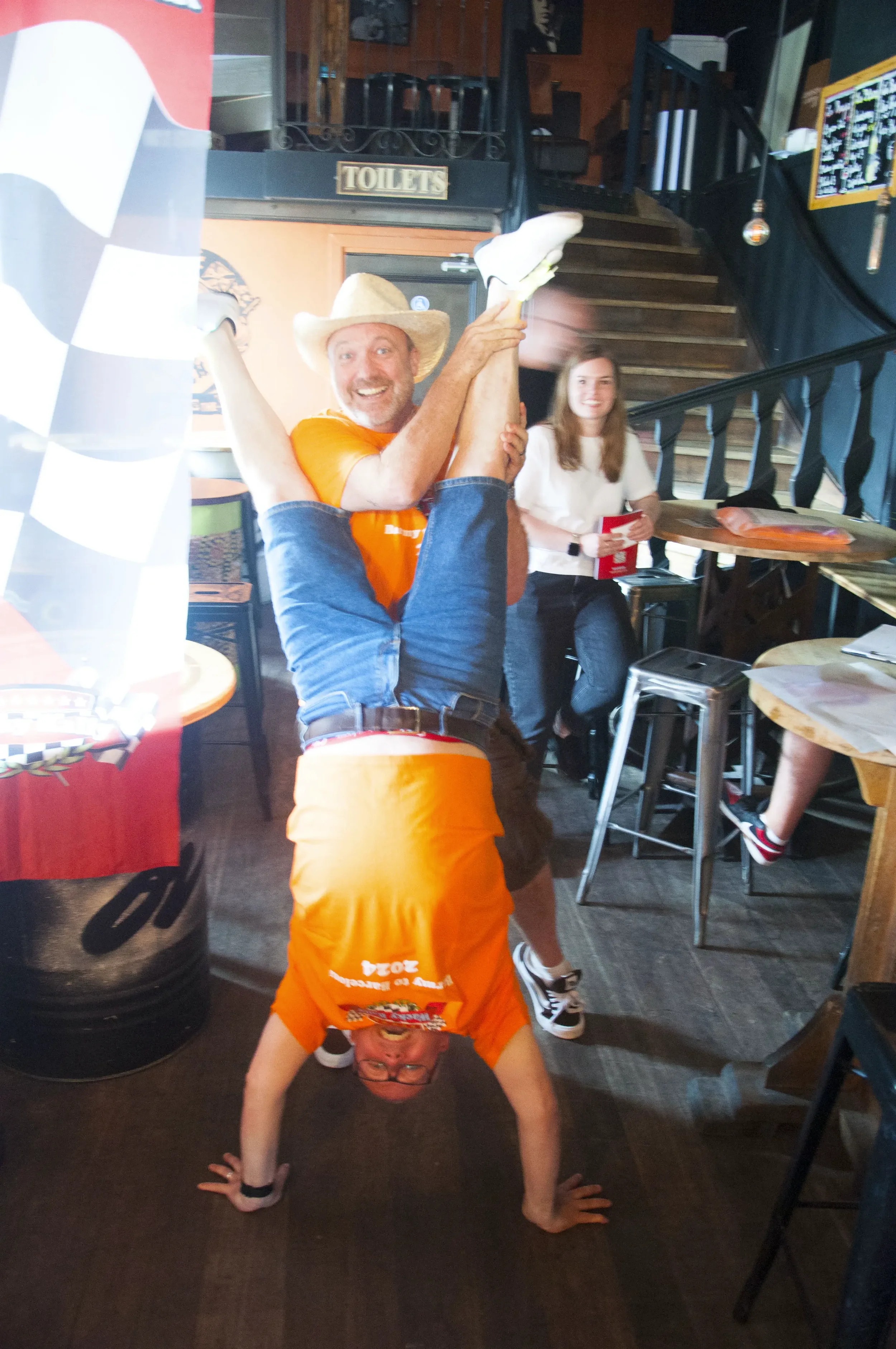 A man wearing a straw cowboy hat and orange shirt being held upside down by another man wearing glasses and a bright orange shirt with the number 2024 on it. They are inside a bar or restaurant.