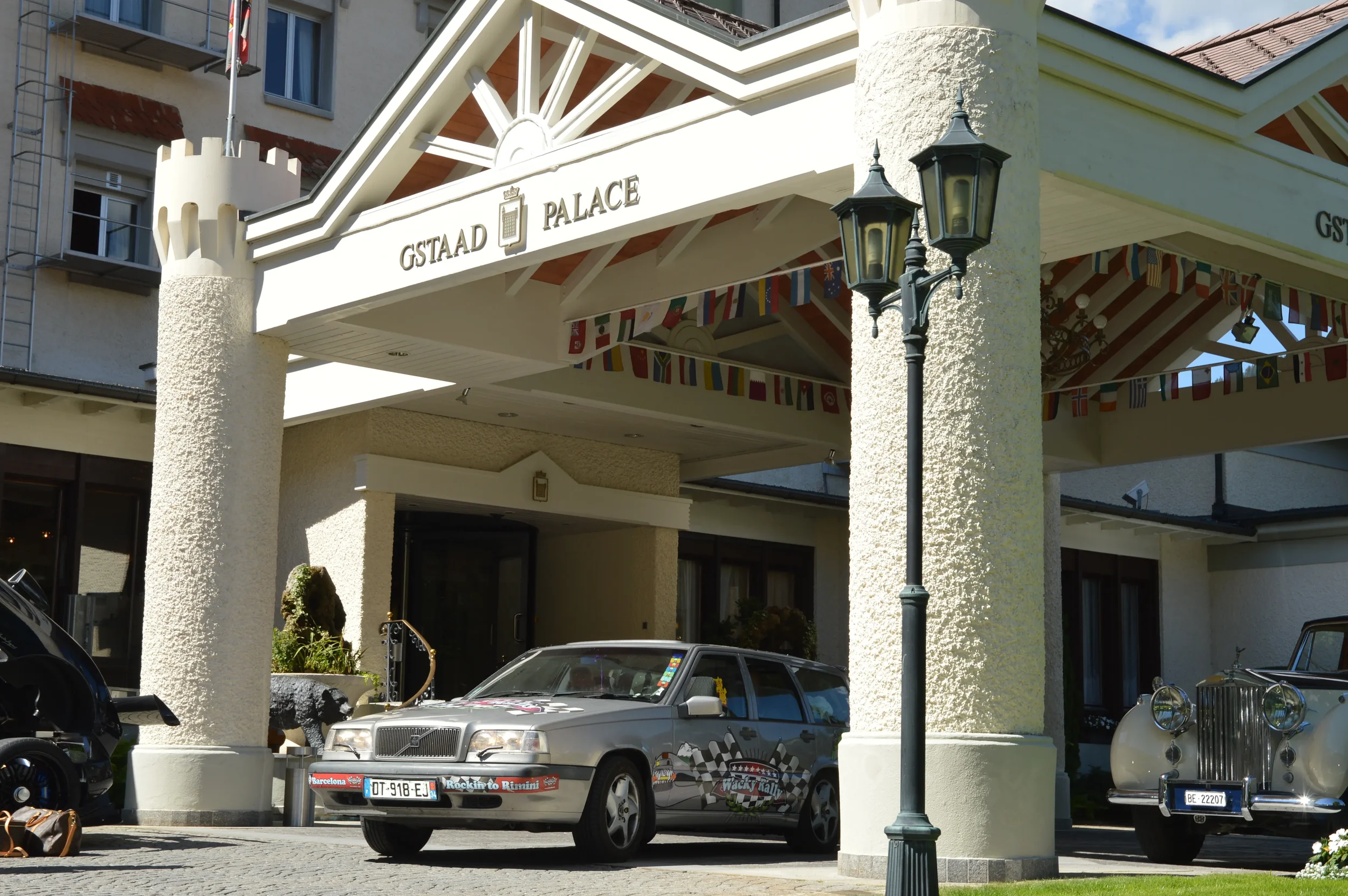 Entrance of Gstaad Palace hotel with vintage cars parked in front, a decorative lamp post, and international flags under the building's awning.