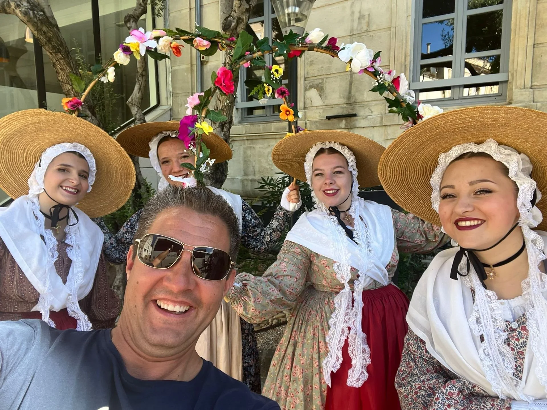 Group of five people, four women dressed in traditional clothing with large straw hats, and one man wearing sunglasses, taking a selfie in front of a building with windows and stone walls. One woman holds a floral arch.