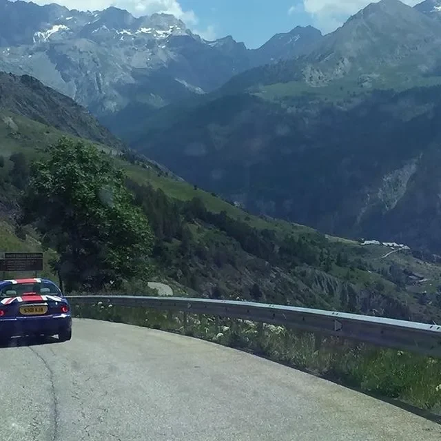 A winding mountain road with a black car, surrounded by green hills, trees, and rugged mountain peaks in the background.