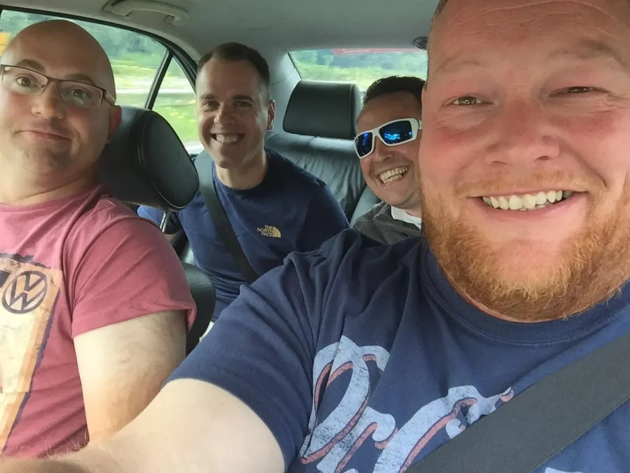 Four smiling men sitting in a car, taking a selfie during a road trip.