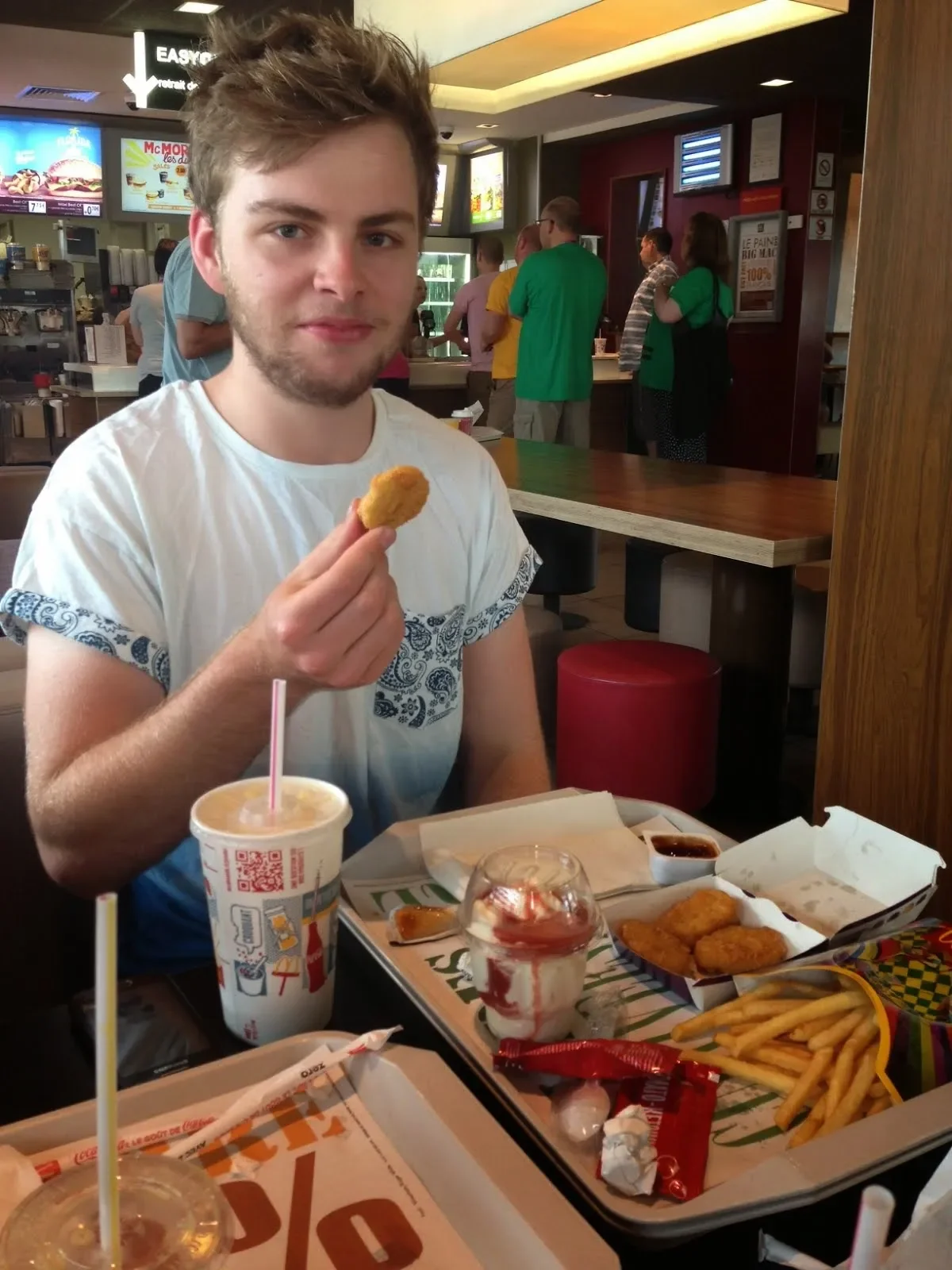 Young man sitting at a table in a fast-food restaurant, holding a piece of chicken, with a tray of fries, chicken nuggets, and drinks in front of him.