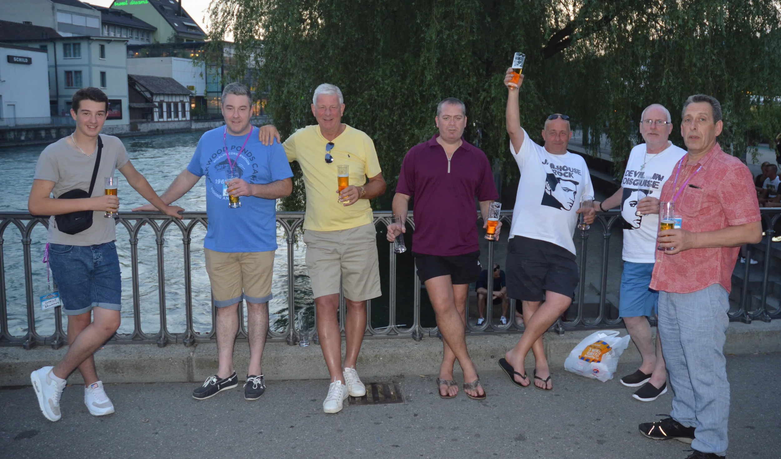 Group of eight men standing by a railing near a river, holding glasses of beer, with trees and buildings in the background during sunset.