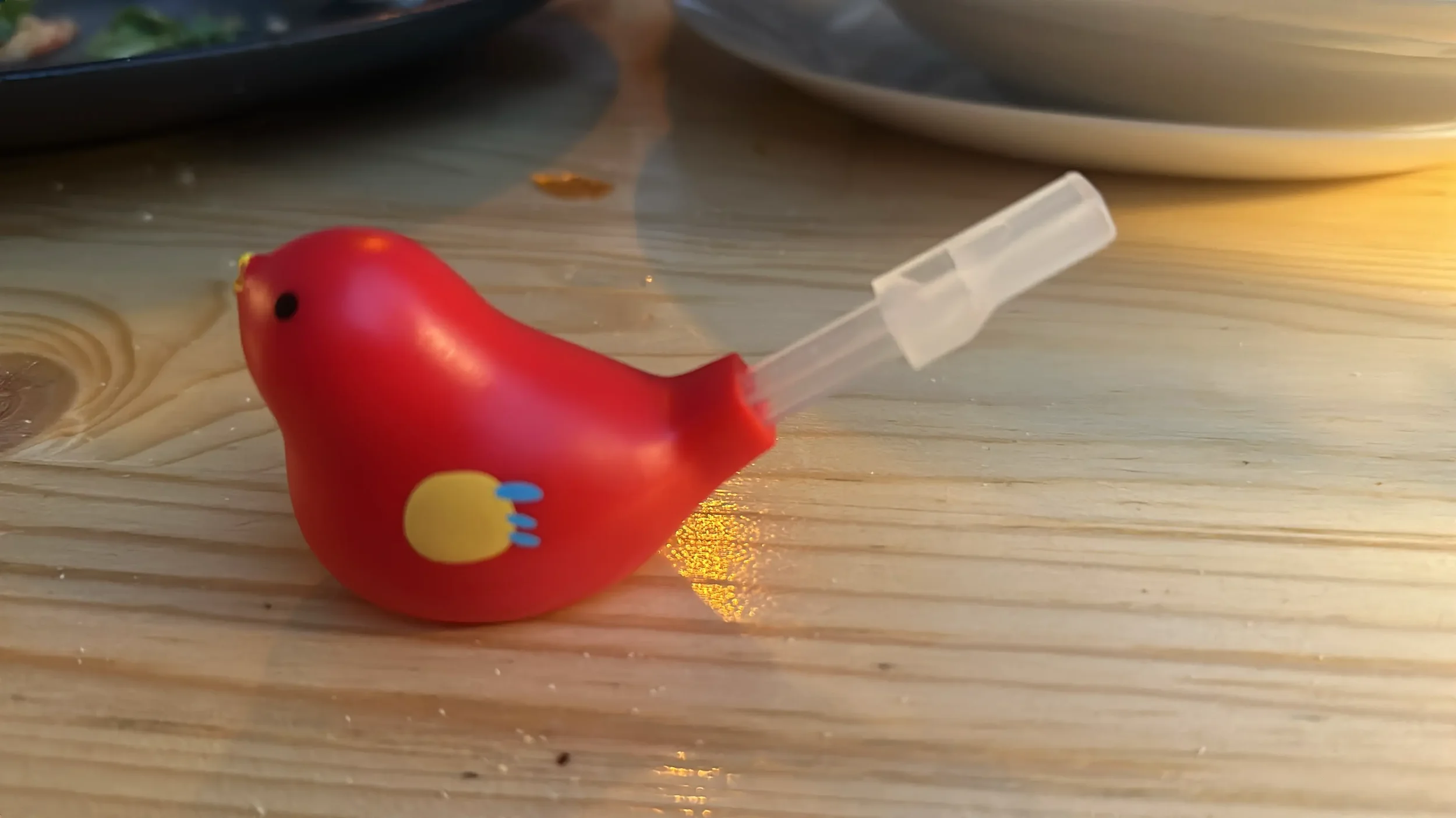 A red bird-shaped object with a yellow wing and black eye, attached to a clear plastic straw, resting on a wooden surface.