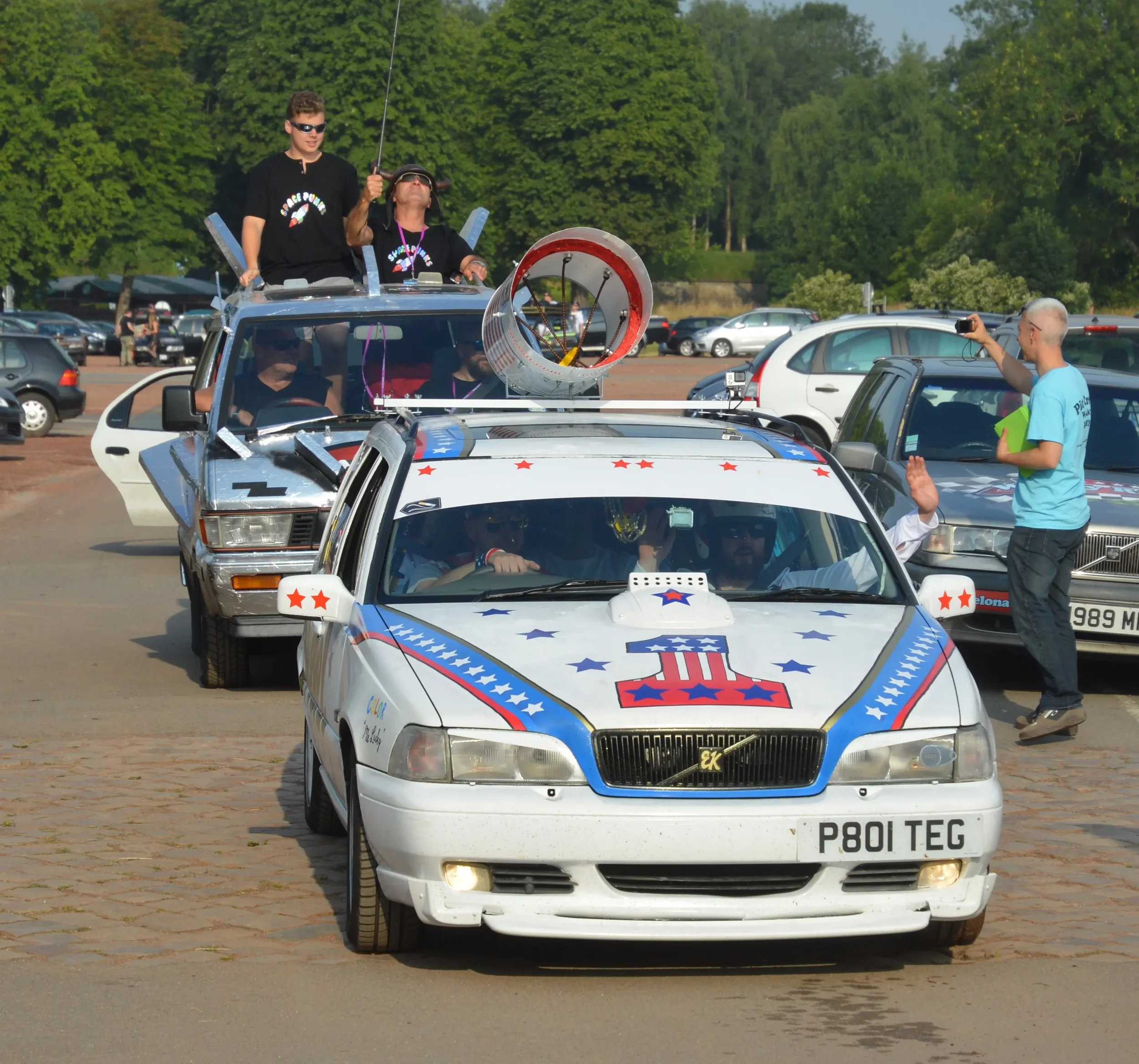 A parade with decorated cars featuring patriotic stars and themes. People are standing and sitting on cars, some taking photos, with trees and parked cars in the background.