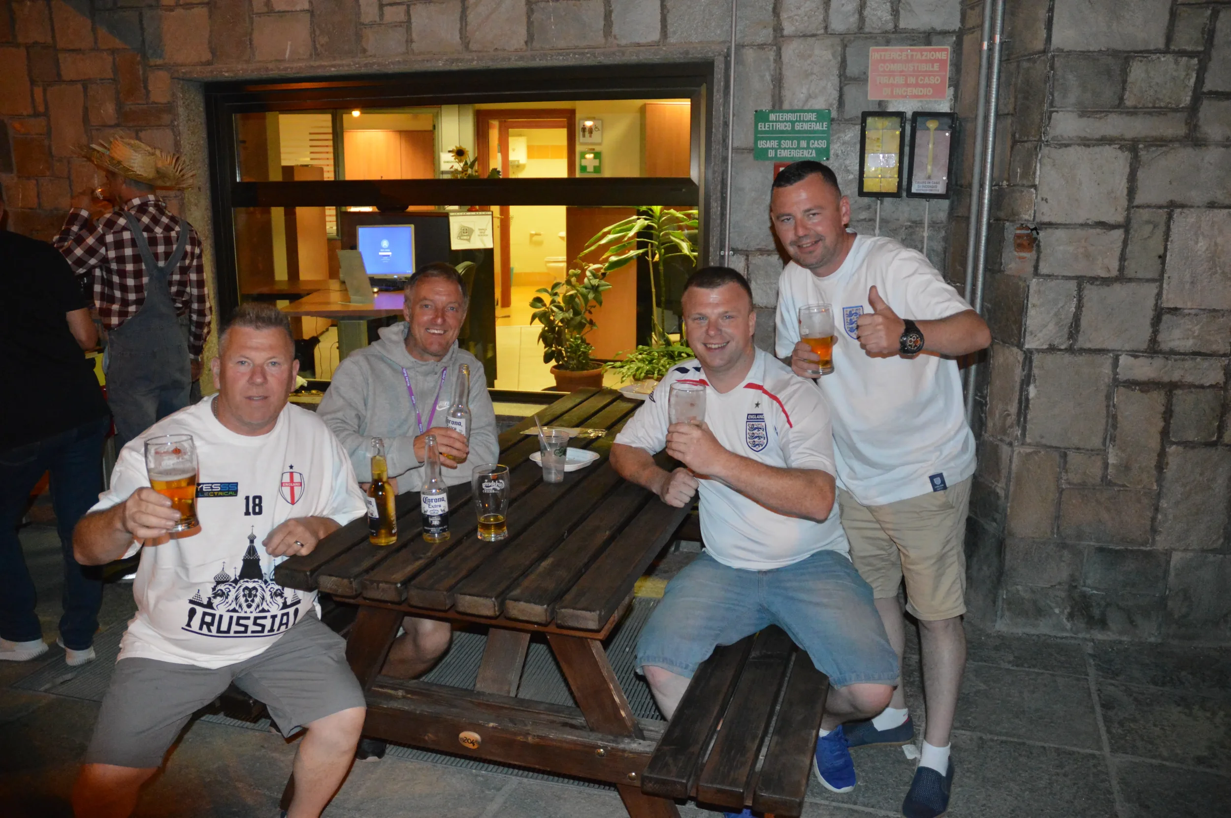 Four men sitting around a wooden table outside, holding glasses of beer, smiling, celebrating, wearing England football shirts, with two of them giving thumbs-up. One man has a Corona beer bottle on the table.