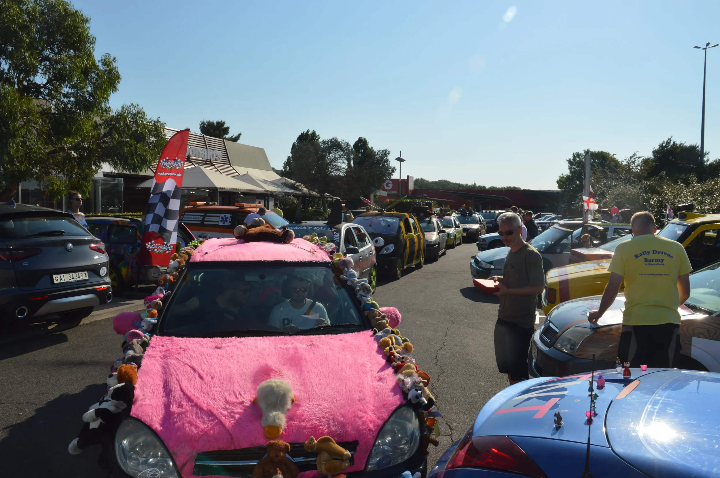 A line of decorated cars at a rally event in a parking lot, with people walking around and inspecting the vehicles during daytime with clear skies.