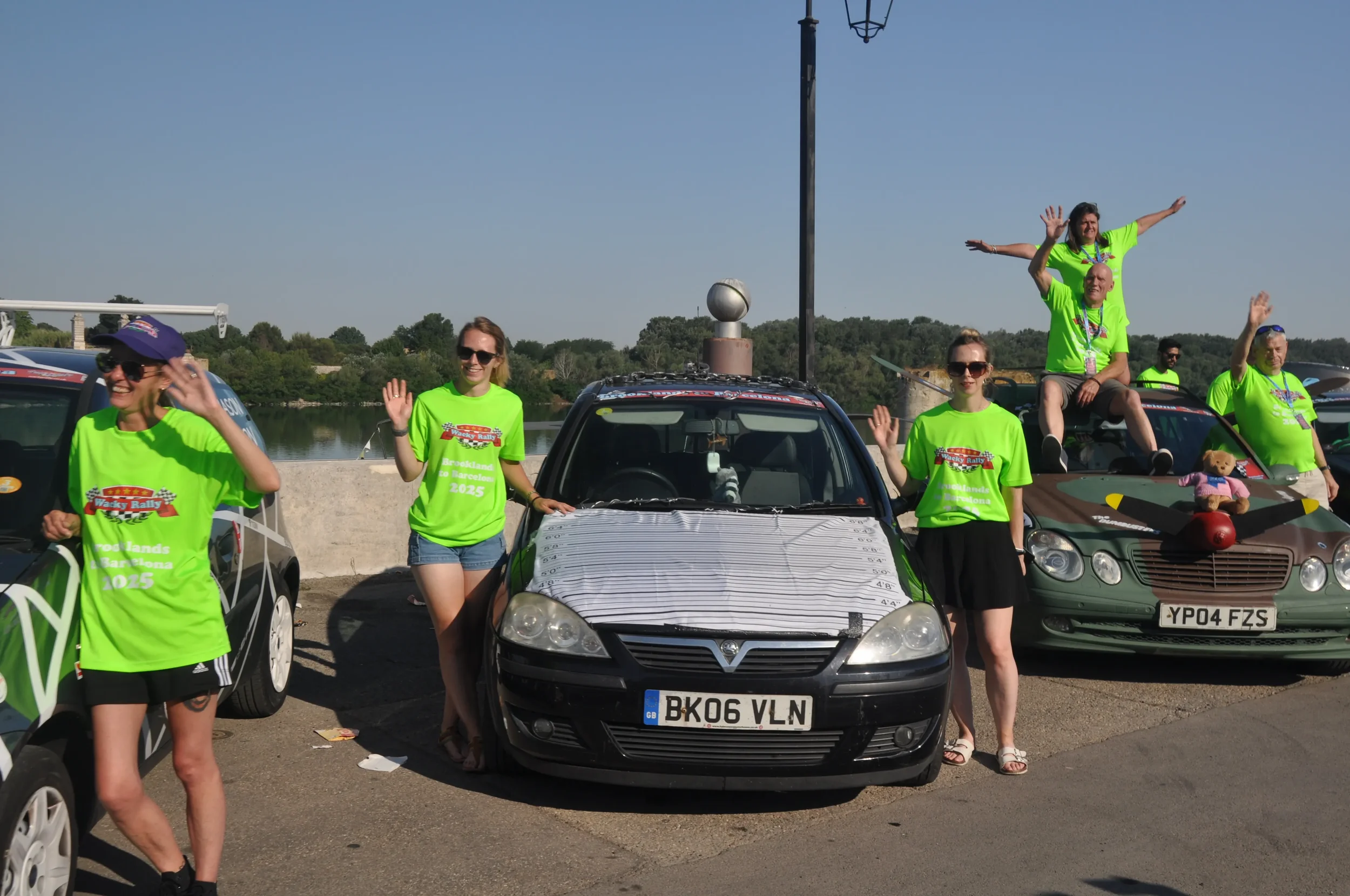 Group of people waving and sitting on decorated cars during outdoor event, possibly a car rally or festival, on a sunny day near a body of water.