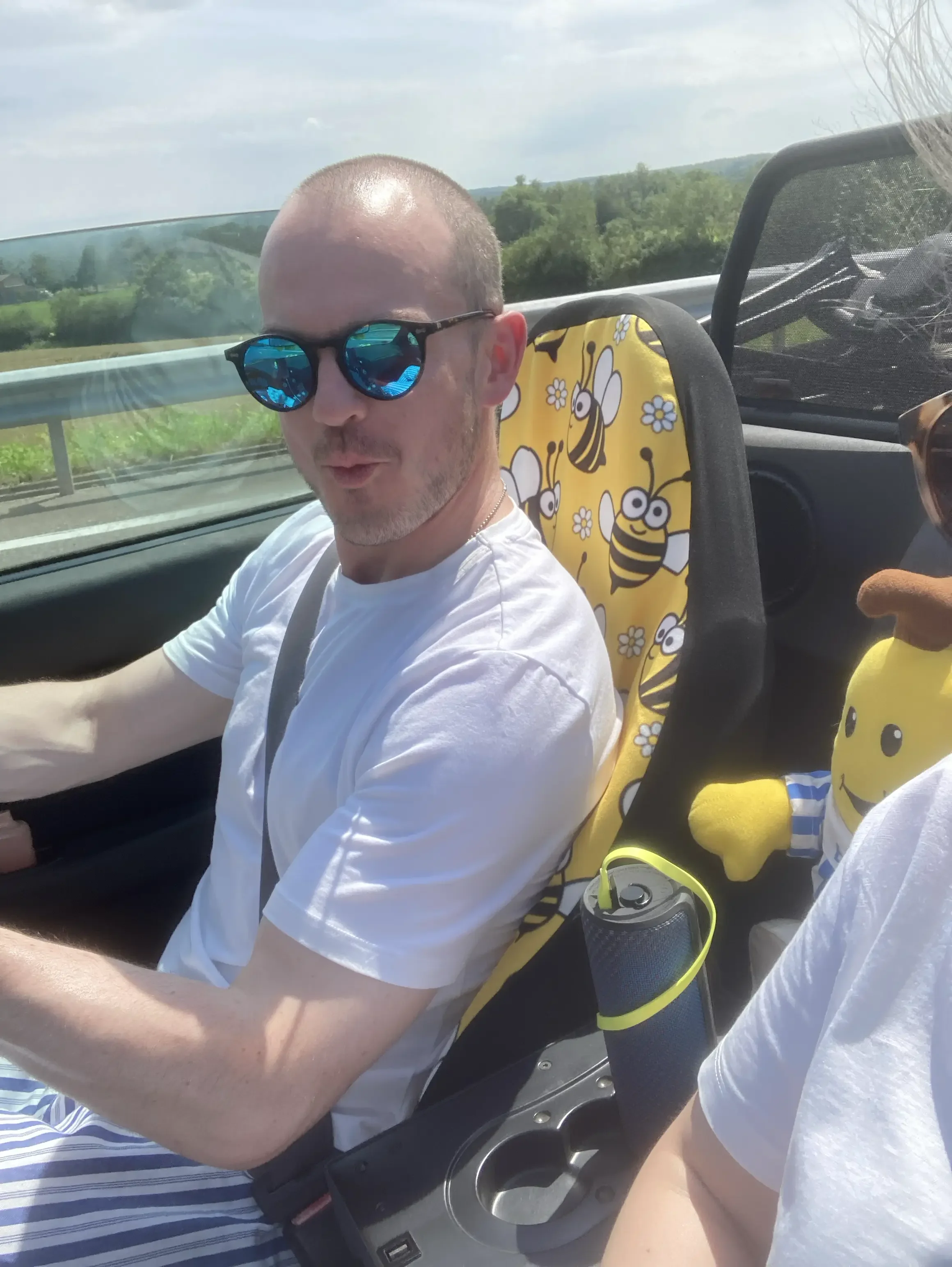 A man with sunglasses driving a convertible with a child in a car seat behind him, countryside scenery visible through the window.