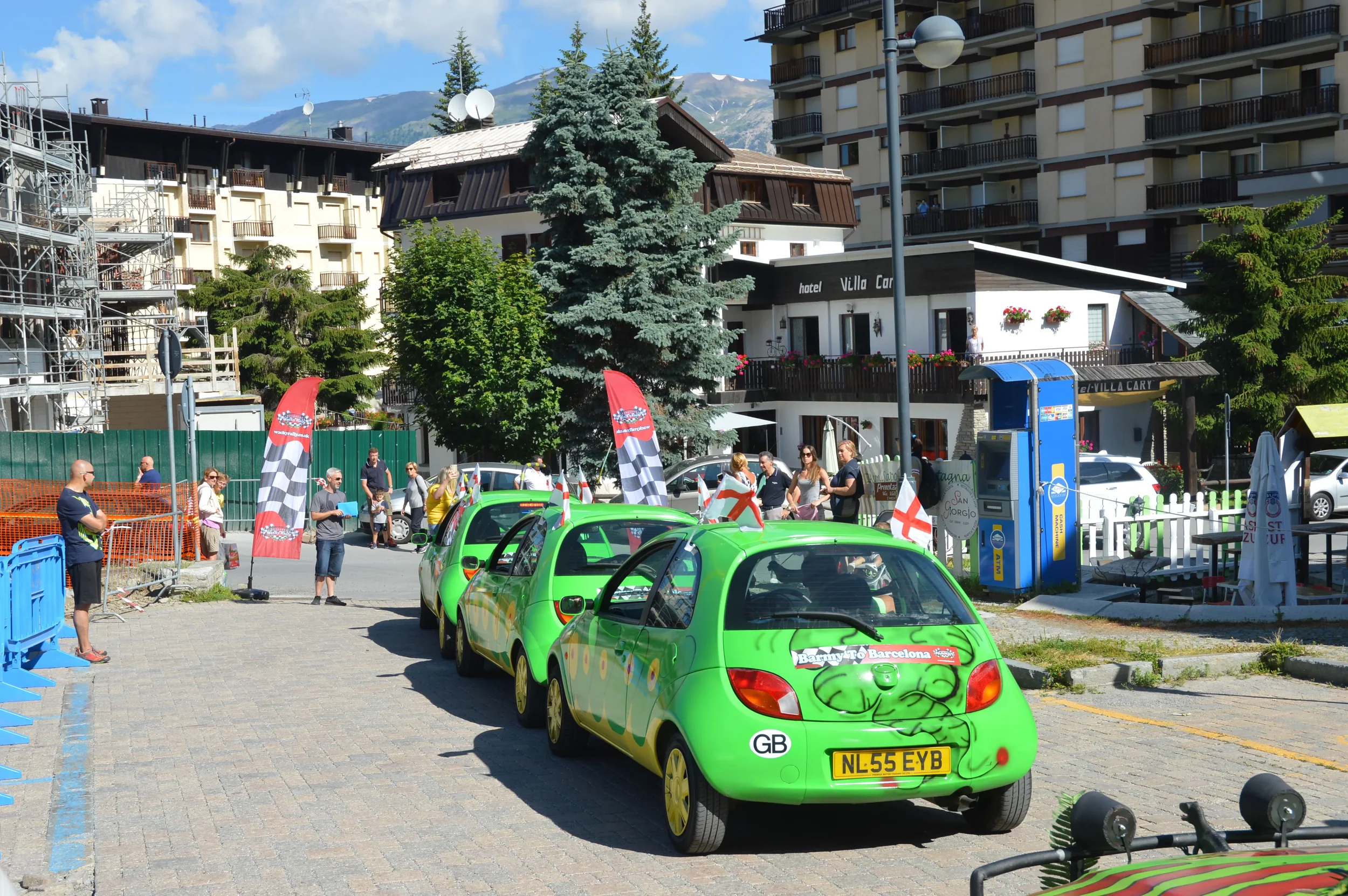A row of four green cartoon-like cars with a frog design on the back, parked in a lot during a daytime event with people walking around and flags with checkered patterns.