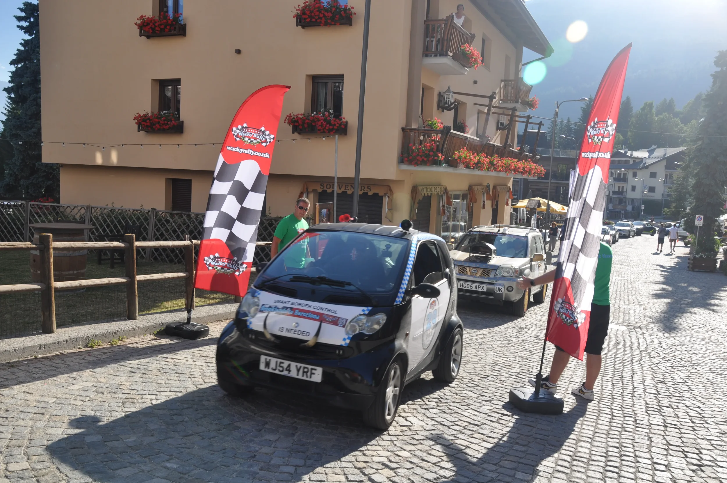 A small electric car with a sign on the front that reads 'Smart Border Control is Needed' is parked on a cobblestone street beside two red flags with checkered patterns and a logo, possibly at a racing or border control event. There are two people ne