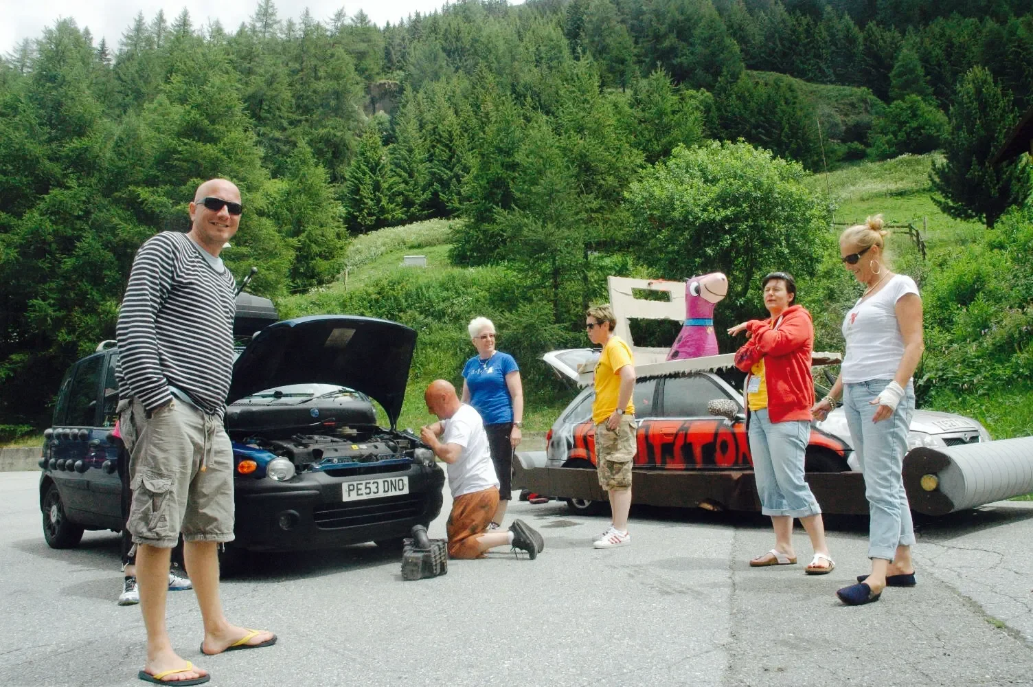 Group of people working on a black car with its hood open on a paved road, with a grassy hillside and trees in the background. One person is kneeling, others are standing, and there is a pink cartoon character on a float attached to a vehicle.