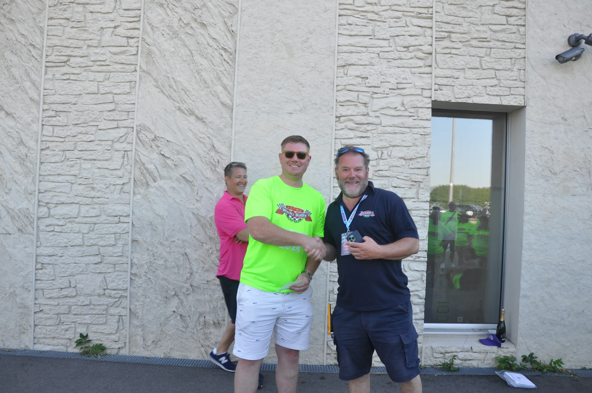 Two men shaking hands outdoors in front of a beige brick wall, smiling at the camera, with a woman in a pink shirt walking behind them and green fields reflected in the window.