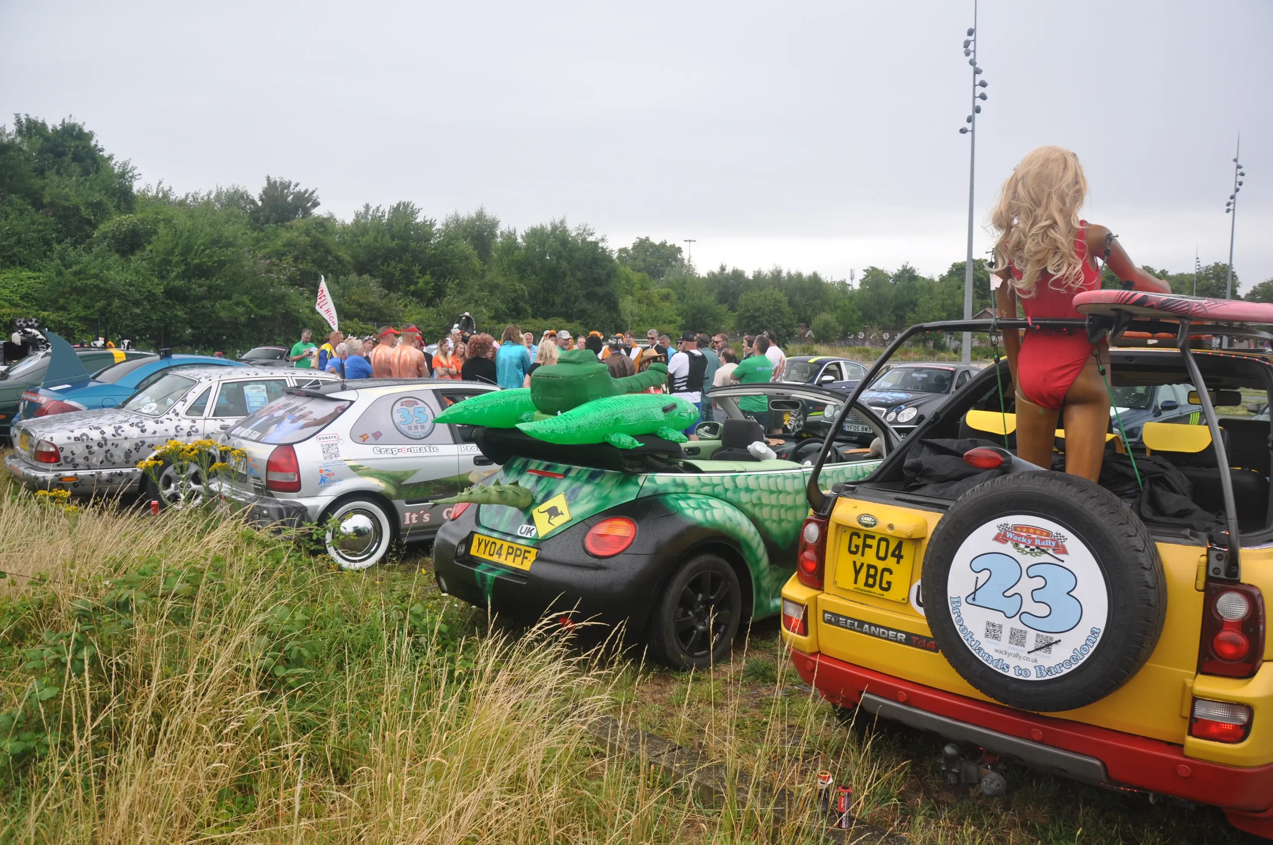 A collection of cars at a gathering, with some vehicles decorated or themed, and a crowd of people in the background outdoors on a cloudy day.